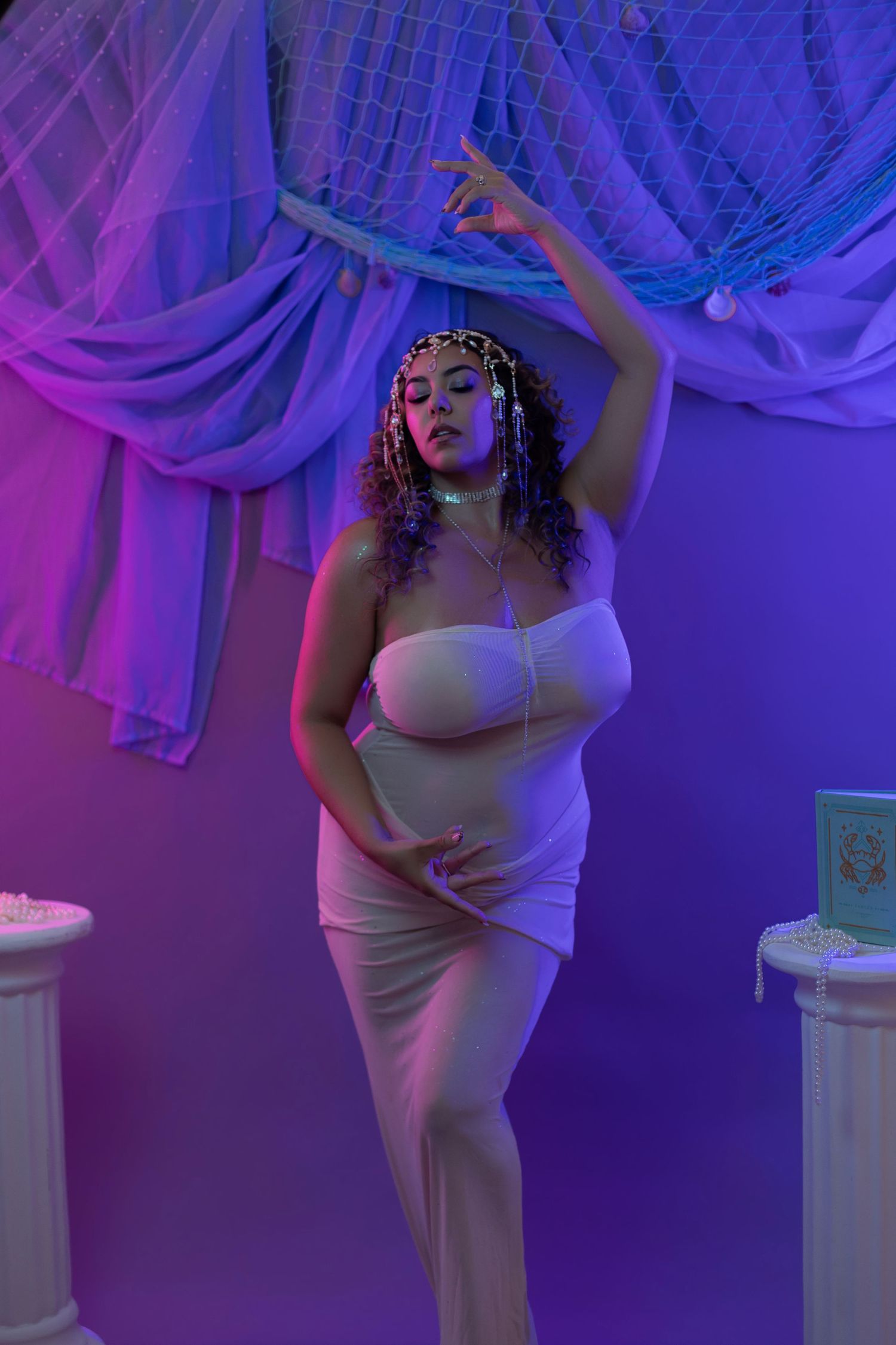 Woman in white strapless dress poses gracefully under purple neon lights with draped fabric backdrop.