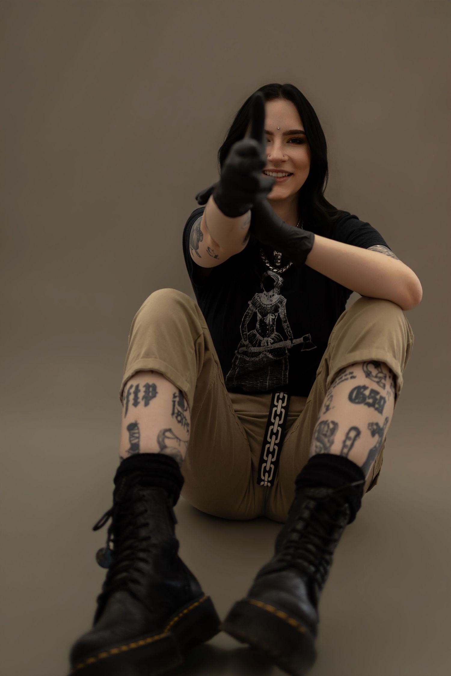 Tattooed woman in black graphic tee, khaki pants, and Doc Martens sits on floor pointing at camera.
