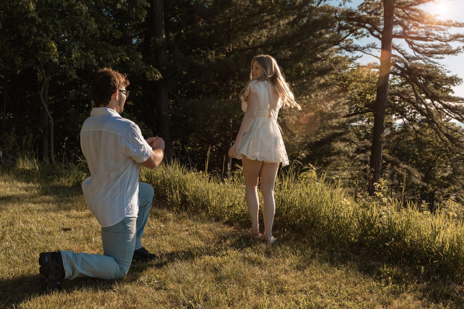 Man on one knee proposing to woman in white dress on sunny grassy hillside surrounded by trees.