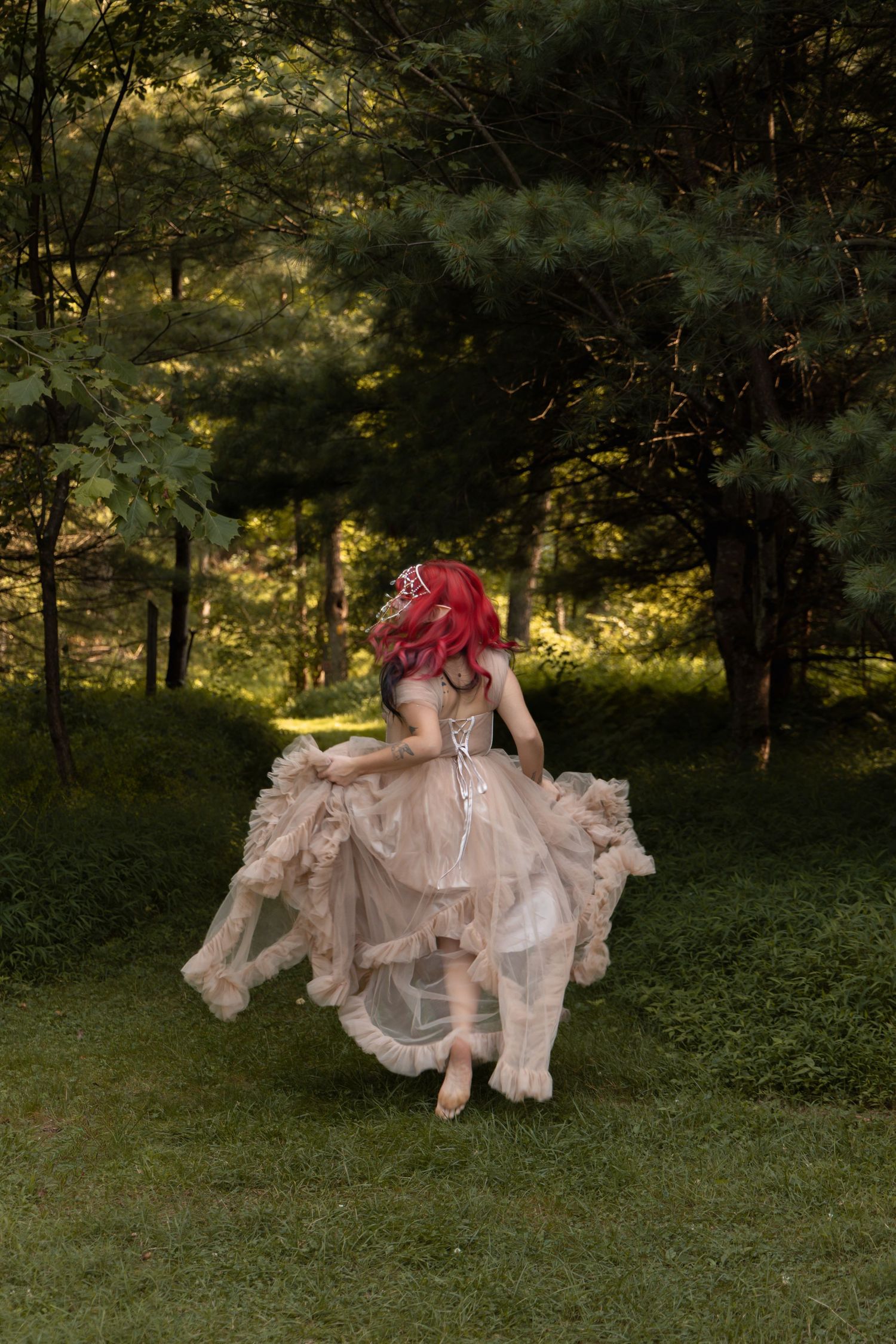 A woman with red hair twirls in a flowing beige tulle gown in a lush green garden setting.