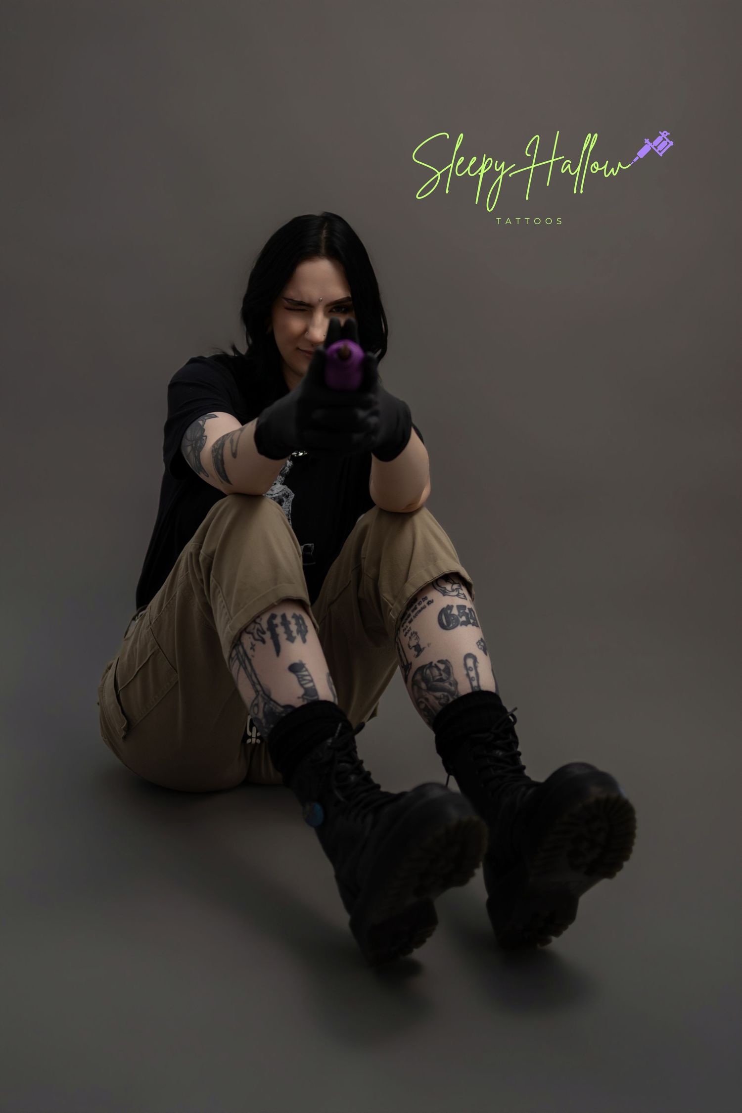 Tattooed woman in khaki pants and black boots sits on floor pointing purple tattoo gun at camera, Sleepy Hallow Tattoos.