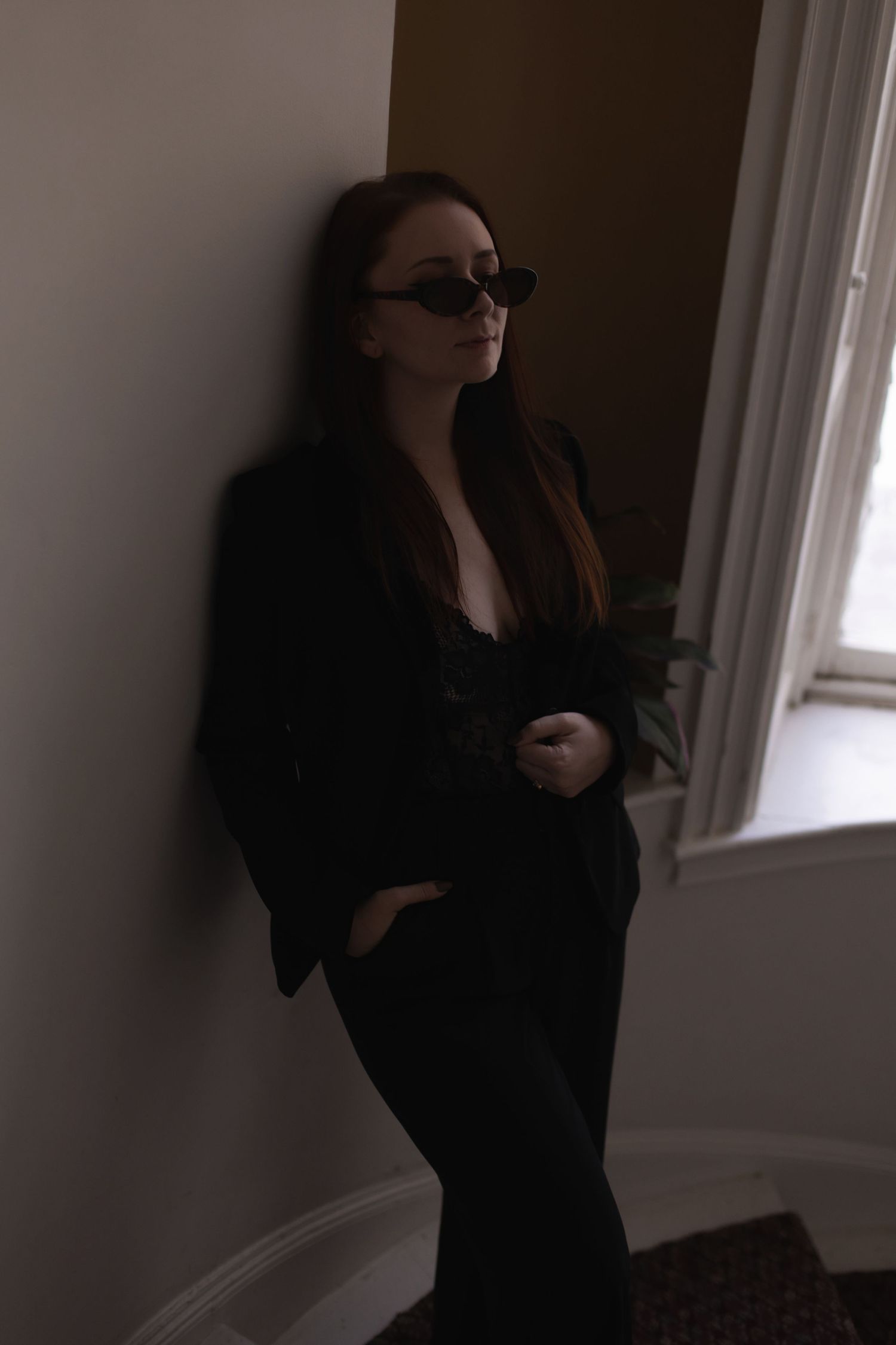 Moody portrait of person in black suit and sunglasses standing by window.