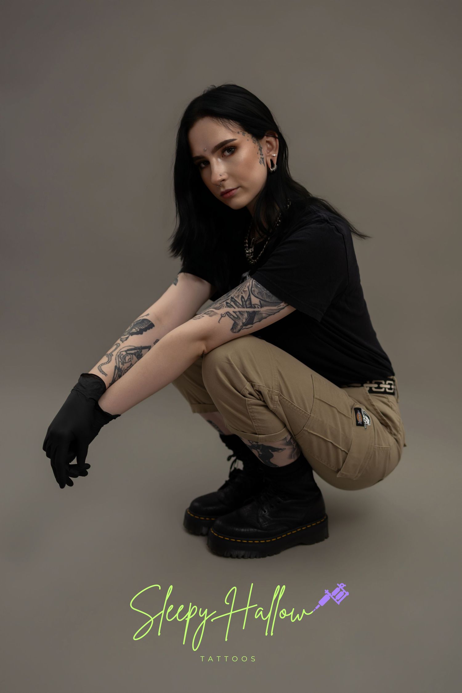 Tattooed female artist crouching in black boots and cargo pants, wearing black gloves, Sleepy Hallow Tattoos logo visible.