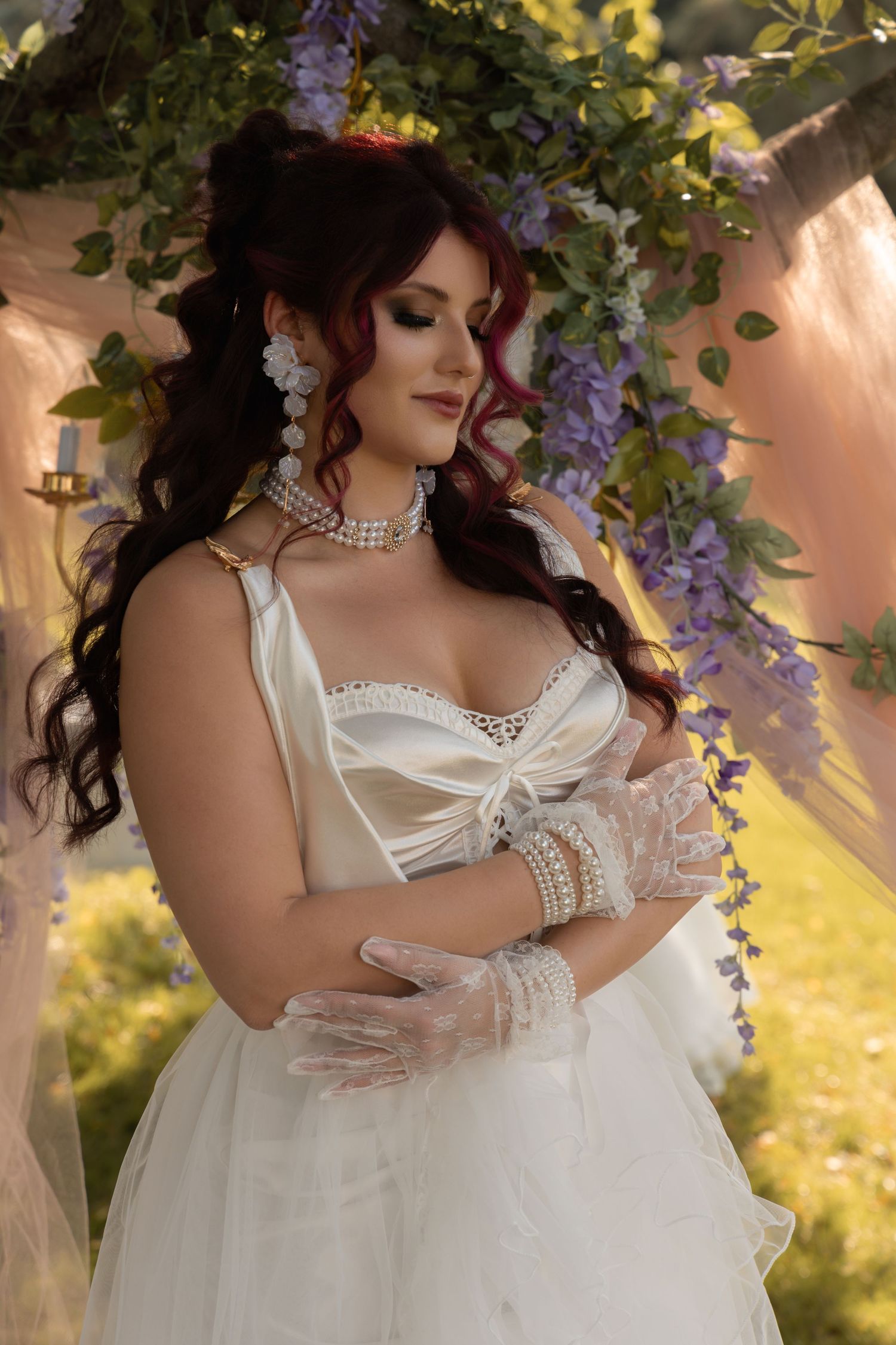 Bride in white satin gown with lace gloves poses under wisteria arch, wearing pearl jewelry and dramatic makeup.