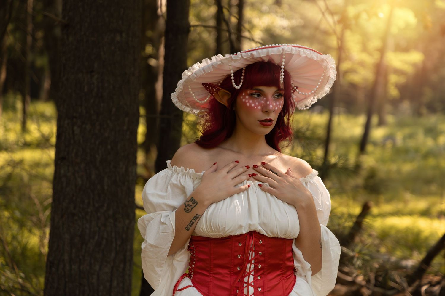 Woman in red corset and white ruffled hat poses in sunlit forest wearing Renaissance costume.
