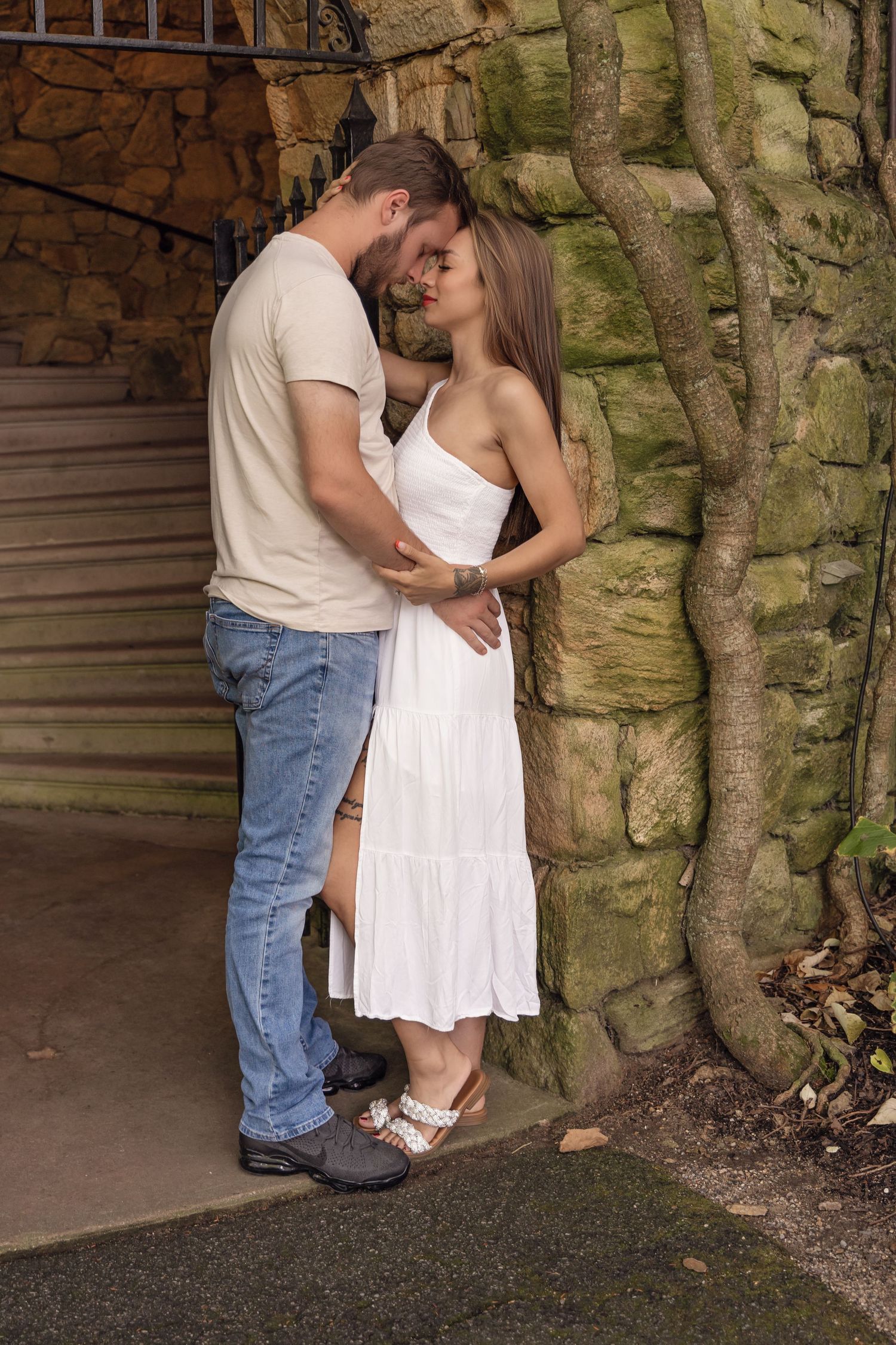 Romantic couple embraces against stone wall in tunnel, with woman in white sundress and man in casual attire.