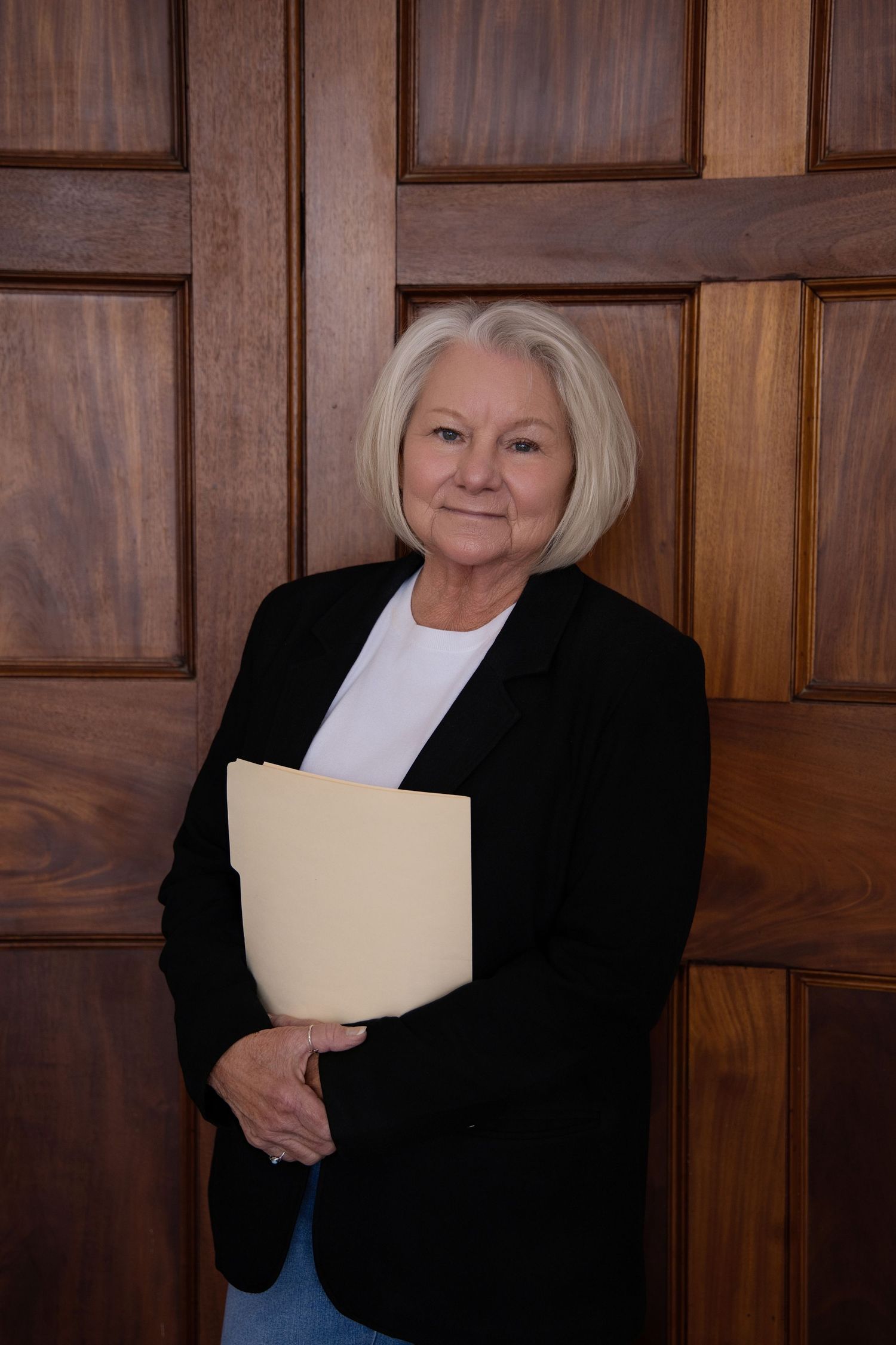 Professional headshot of someone in business attire holding a folder against wood paneled wall.