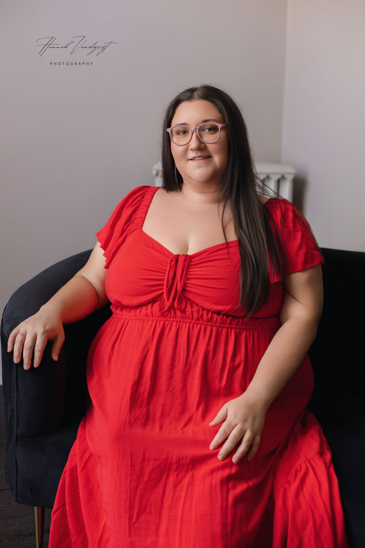 Woman in vibrant red dress poses confidently on dark couch during indoor photoshoot.