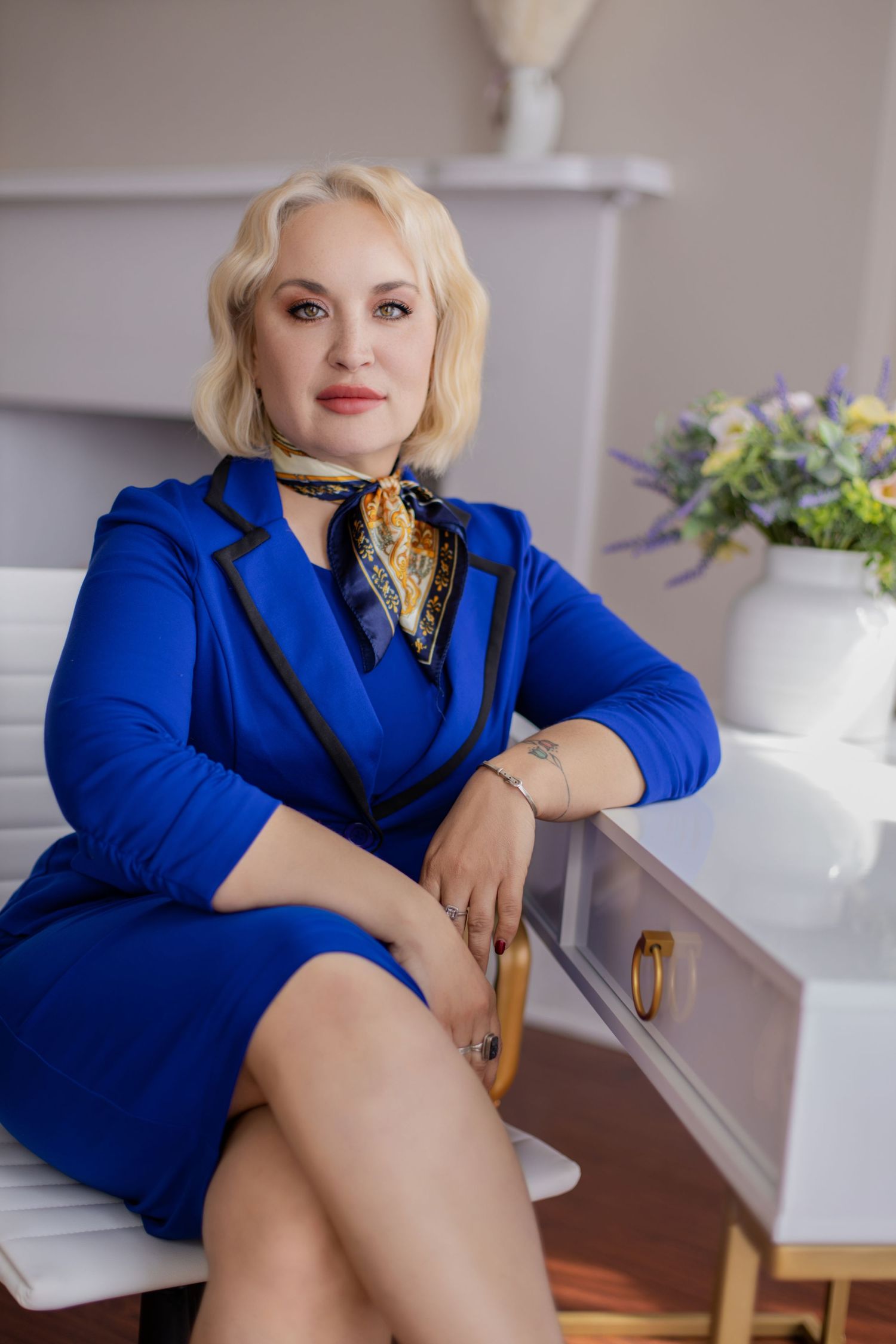 Sophisticated pose in bright blue blazer with silk scarf near white floral arrangement.