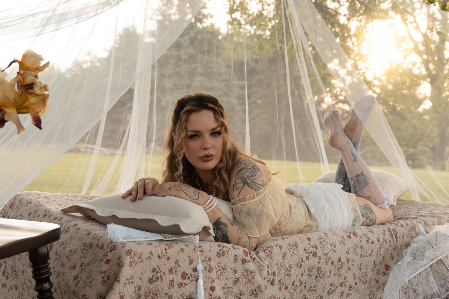 Tattooed woman lying on a floral bed outdoors under sheer canopy draping, bathed in warm golden sunlight.