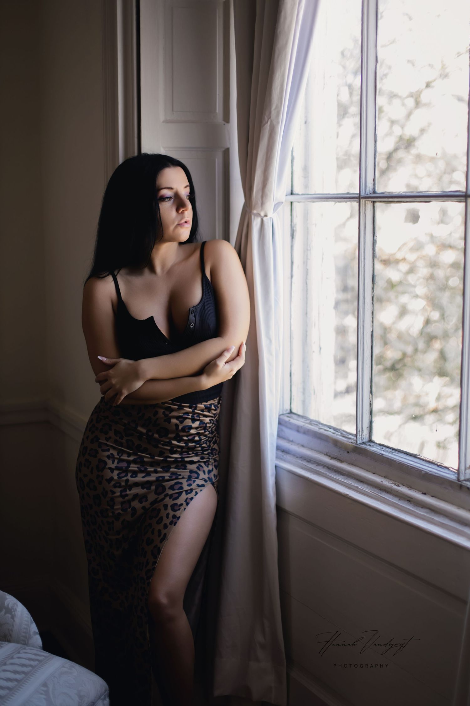 Model in black top and leopard print skirt poses by window in moody lighting.