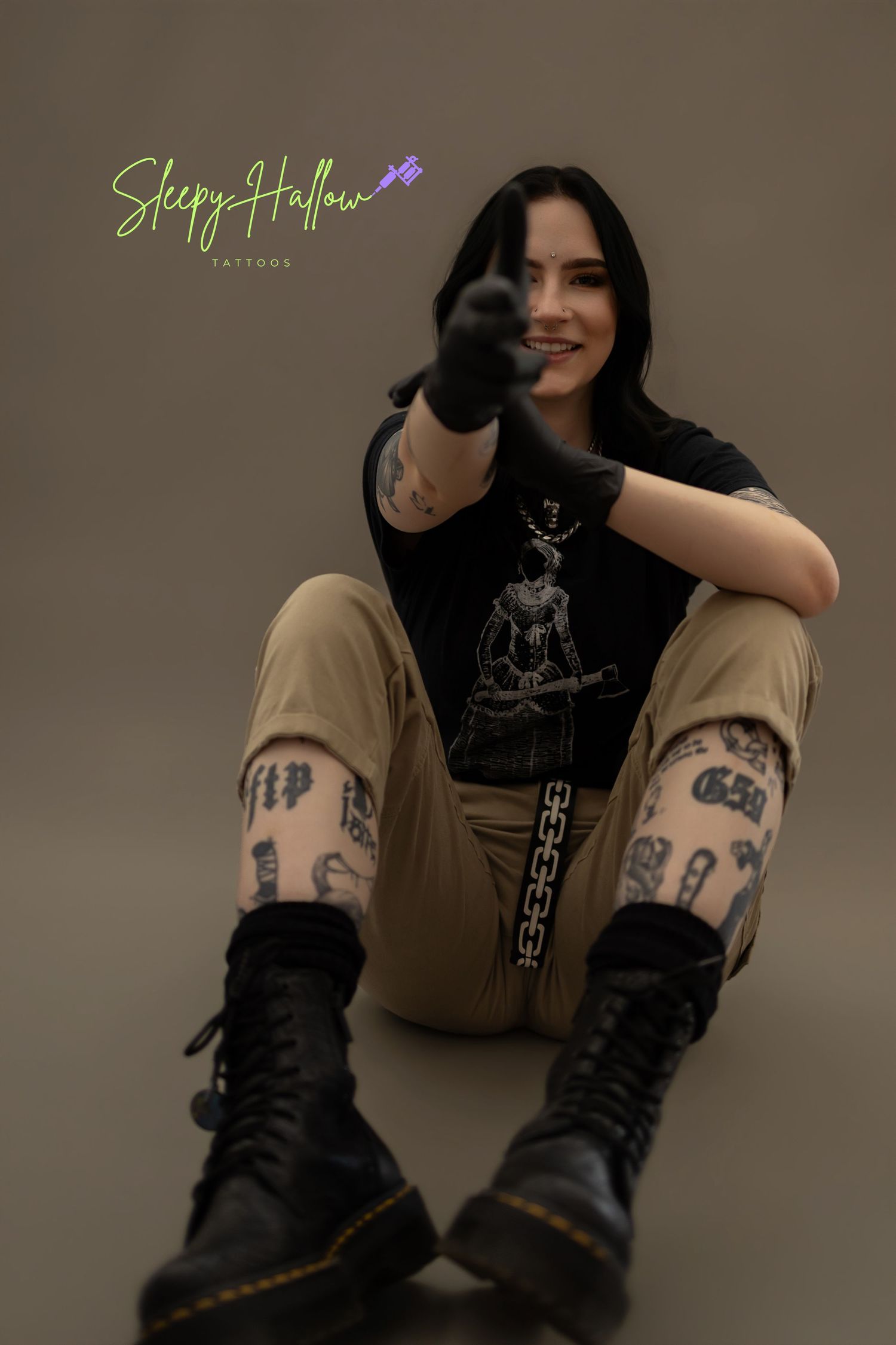 Tattooed woman in black graphic tee, khaki pants, and Doc Martens sits on floor pointing at camera, smiling.
