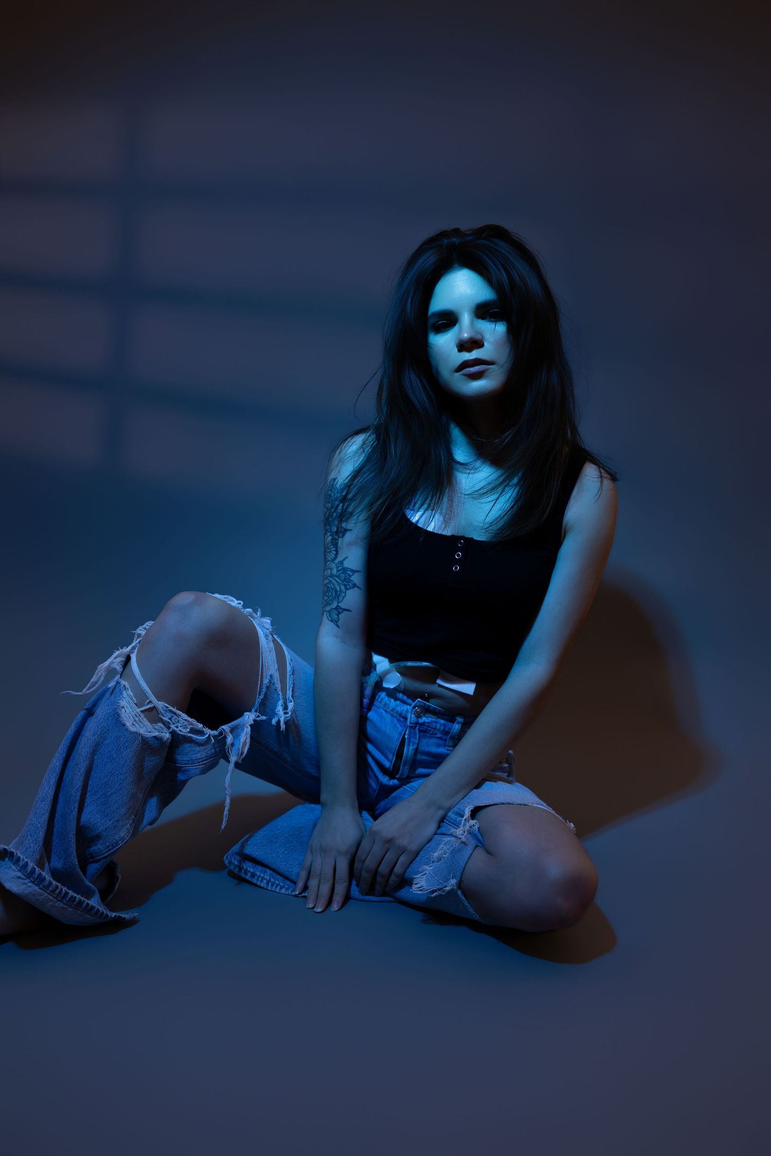 Young woman with dark hair and tattoo poses on floor in ripped denim shorts and black top under blue studio lighting.