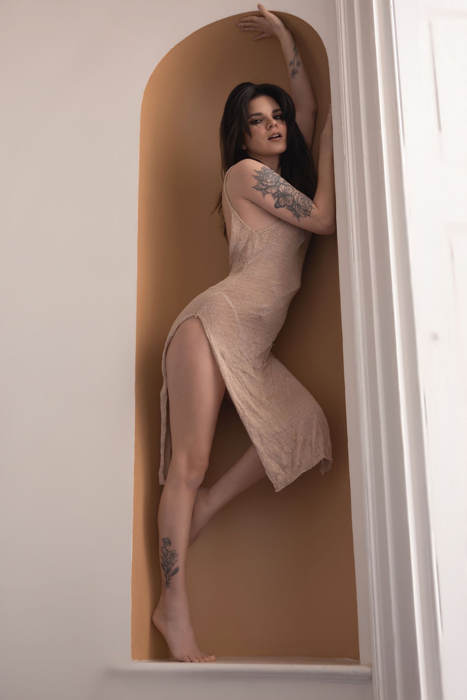 Woman in beige slit dress with tattoos posing elegantly in an arched alcove.