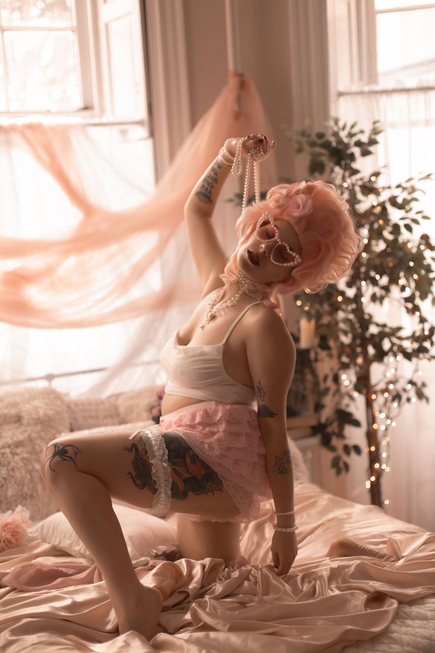 Pink-haired woman with tattoos poses on a bed in white bralette and pink skirt, holding pearls in a dreamy room.