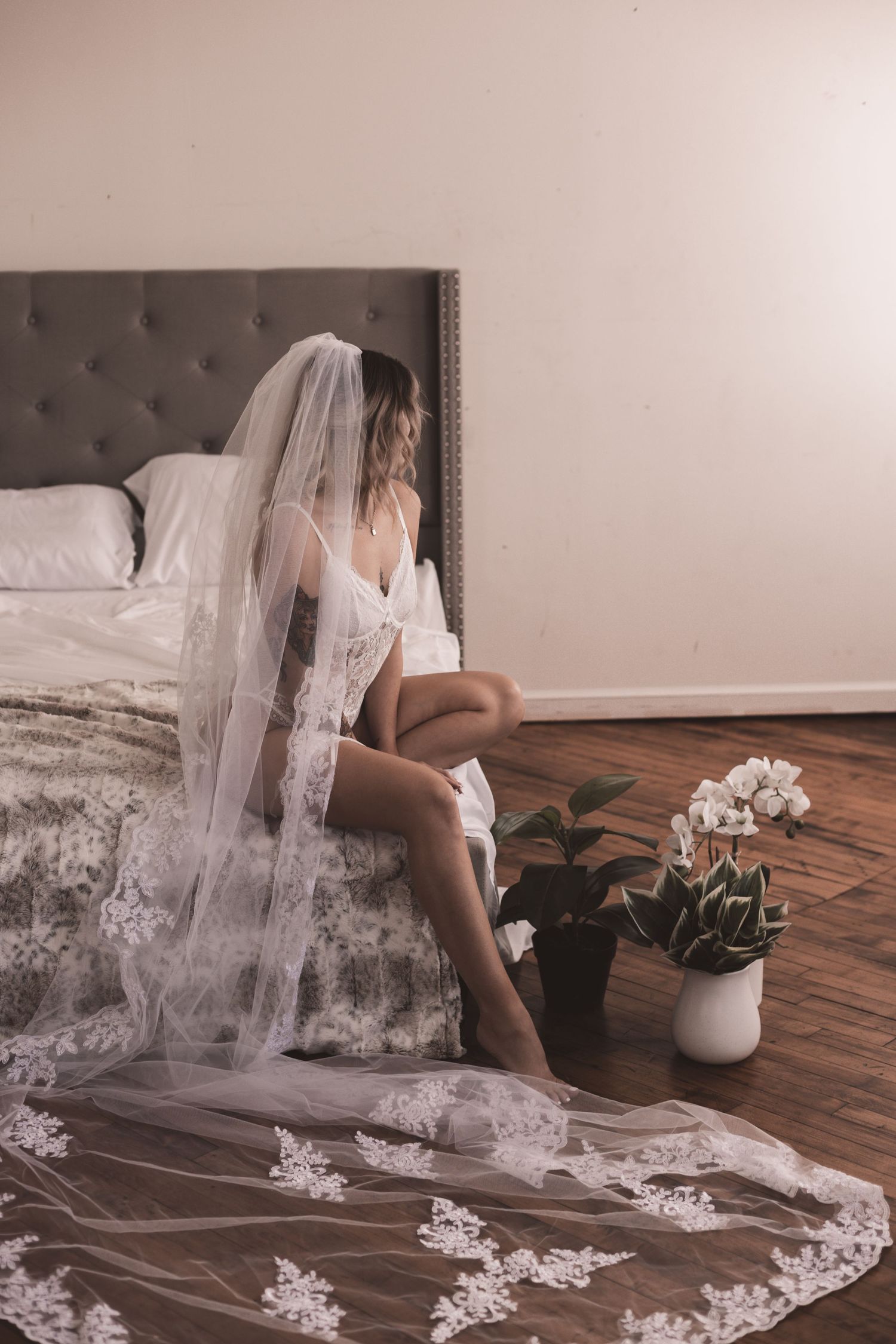 Woman in bridal lingerie sits on grey tufted bed with lace veil flowing to floor.