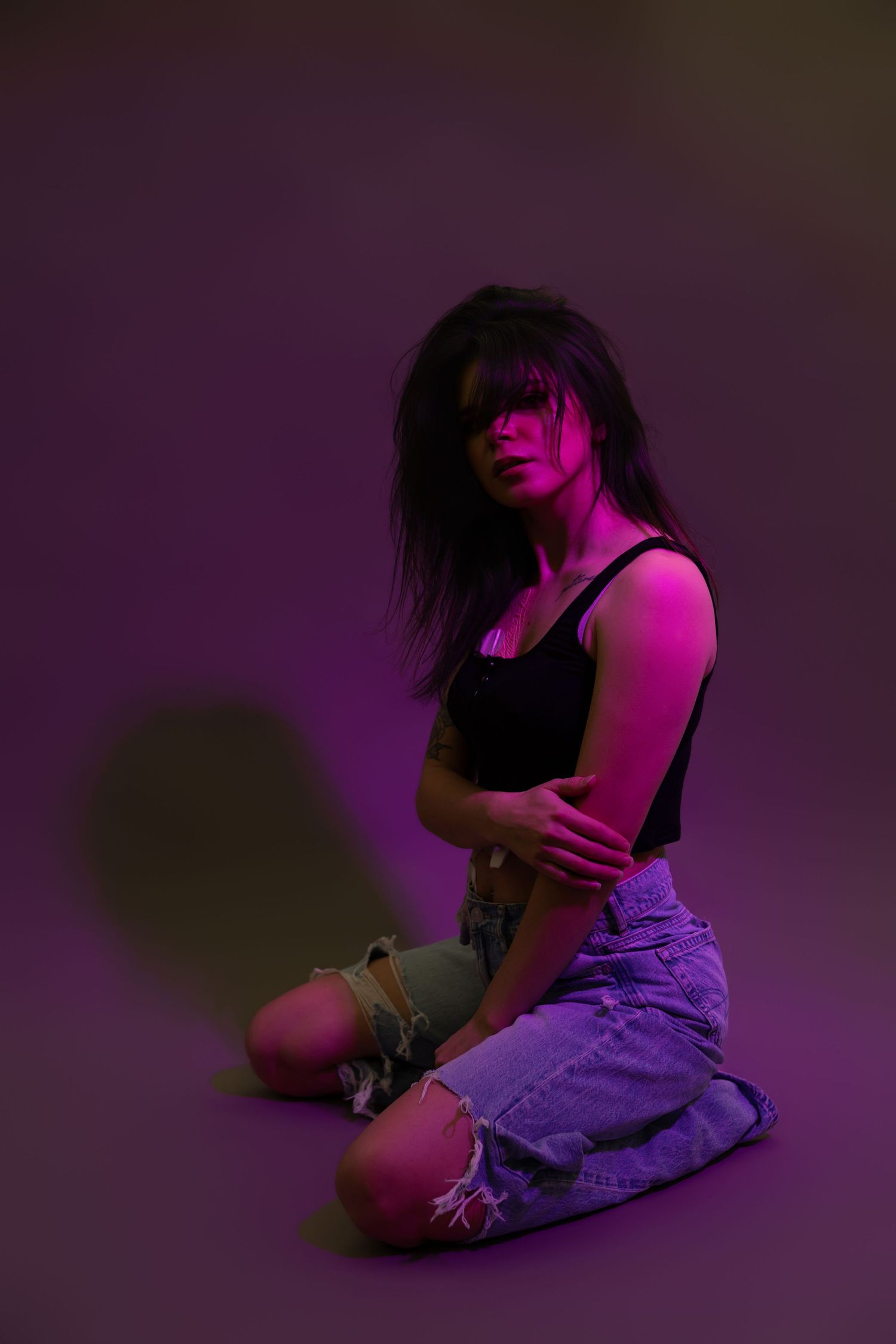 Woman in black crop top and ripped jeans sitting on floor under purple and pink neon studio lighting.