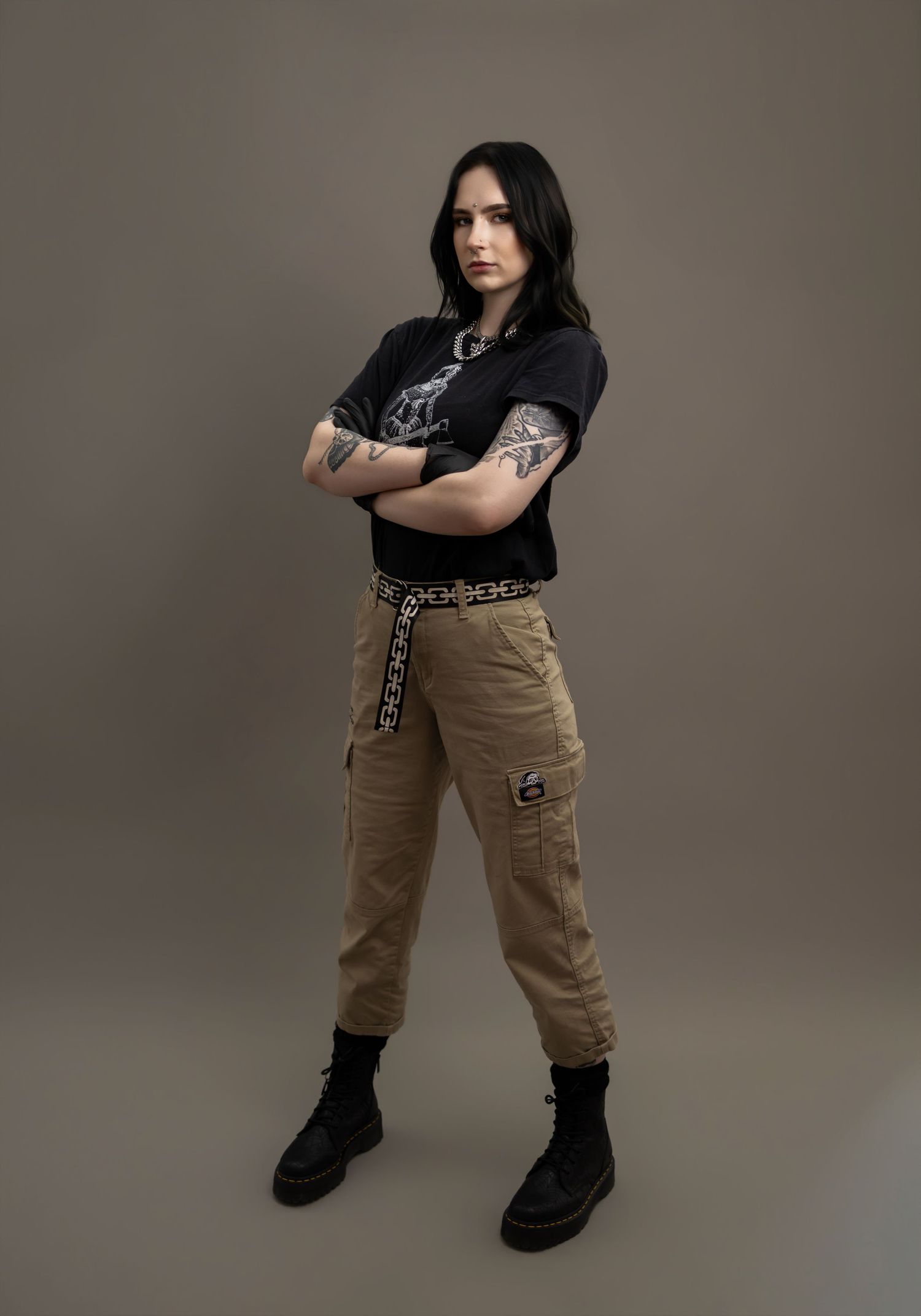 Tattooed woman standing with arms crossed in black graphic tee, khaki cargo pants, and black boots.