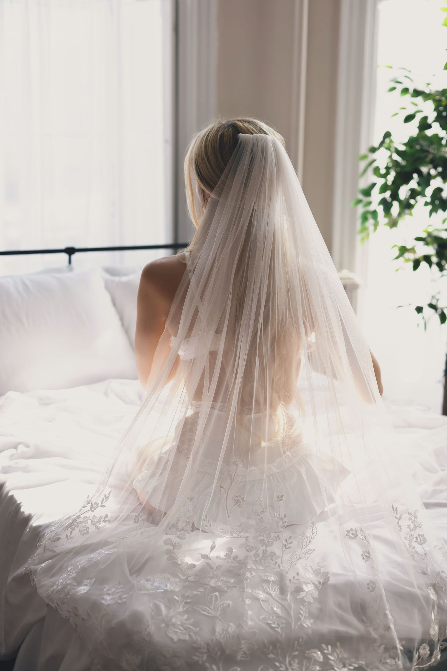 Bride in white lace gown and veil sitting on bed, viewed from behind near bright window.