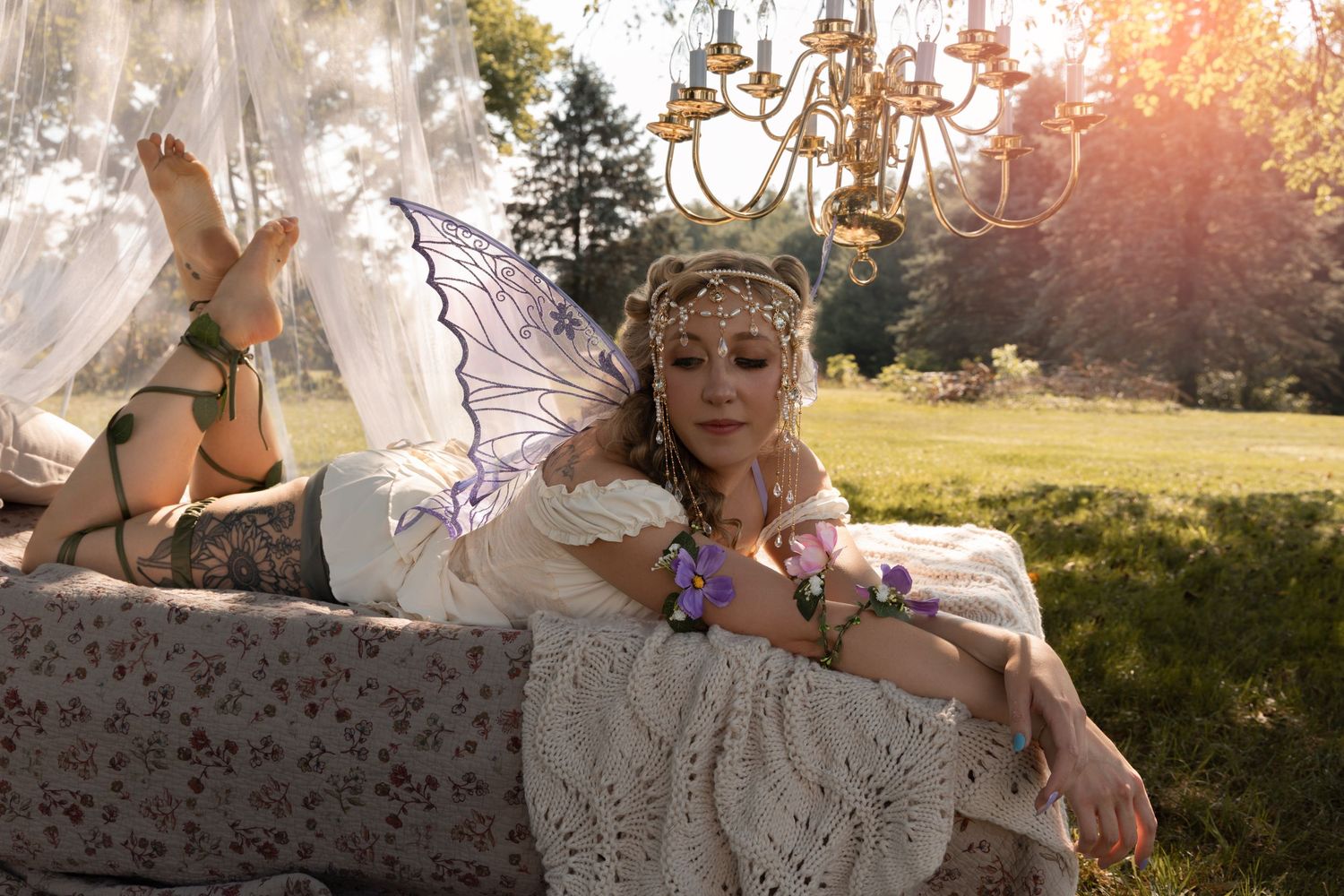 A woman in fairy costume with wings and floral crown reclines outdoors near a chandelier in golden sunlight.