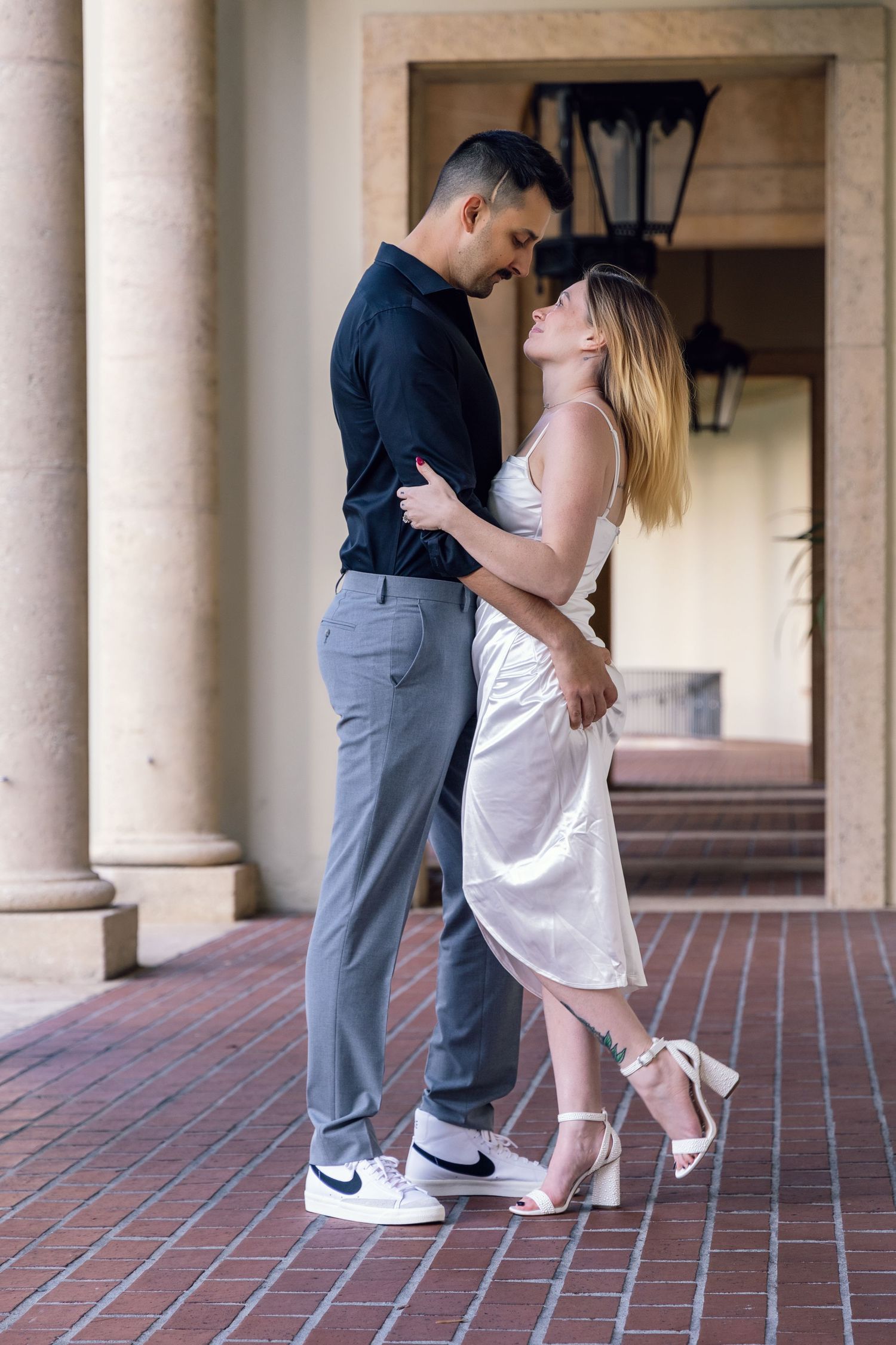 A romantic couple embraces on a brick patio, with her in a white dress and him in a navy shirt and grey pants.