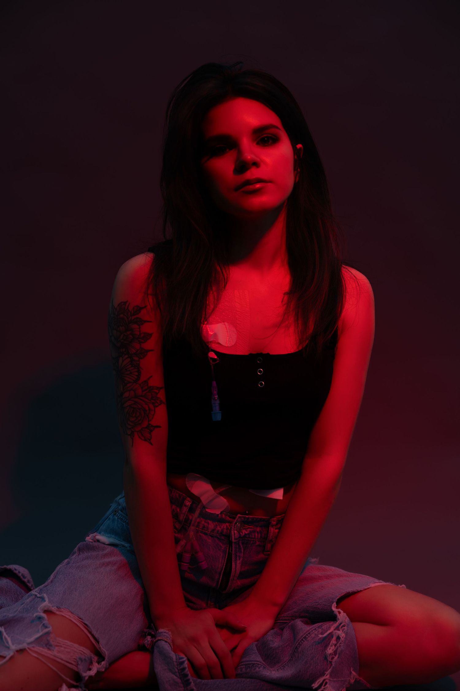 Young woman with tattoo posing in red neon light wearing black crop top and ripped denim shorts.