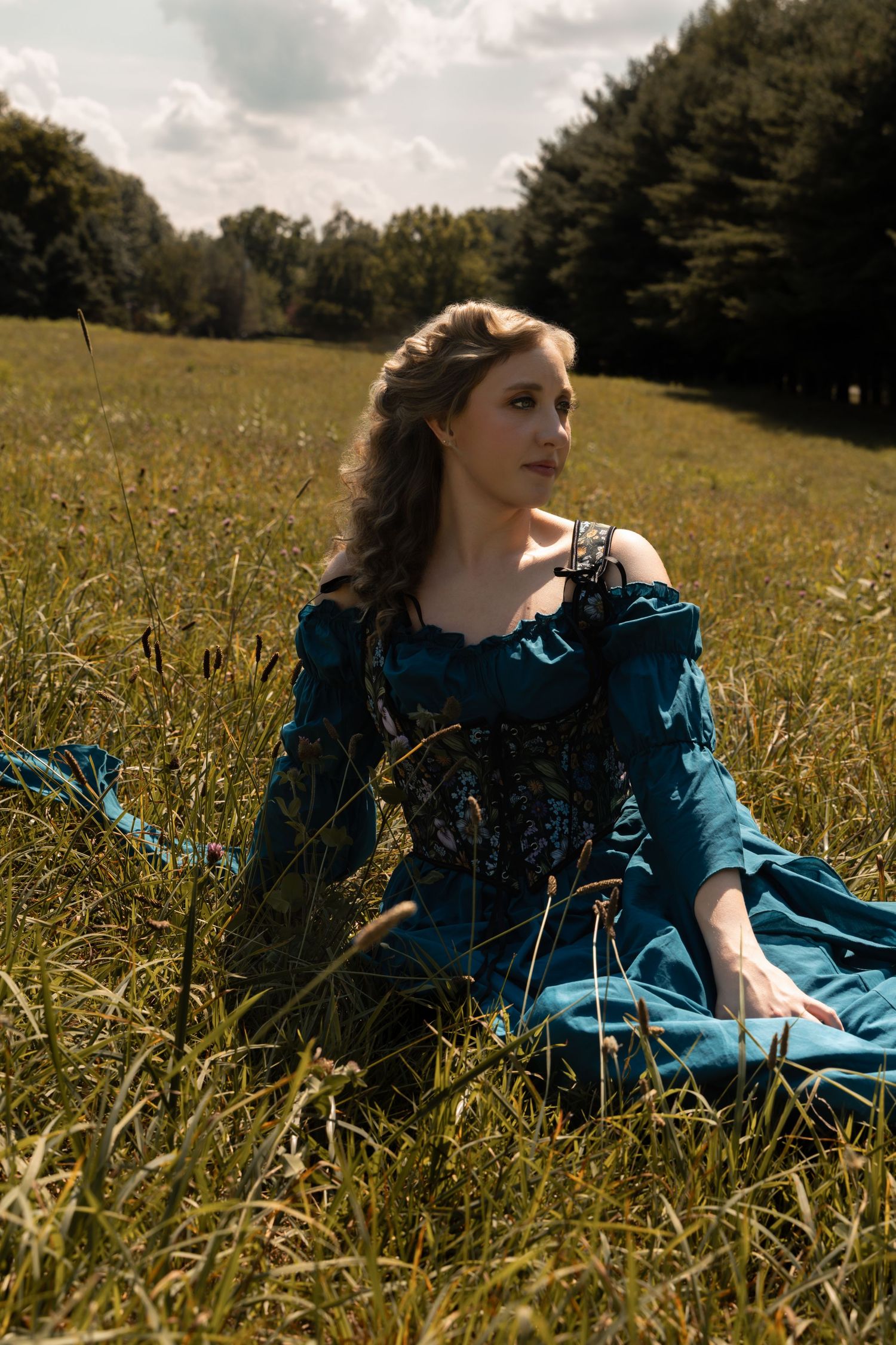 Woman in teal medieval gown sitting gracefully in a sunlit green meadow with trees in background.