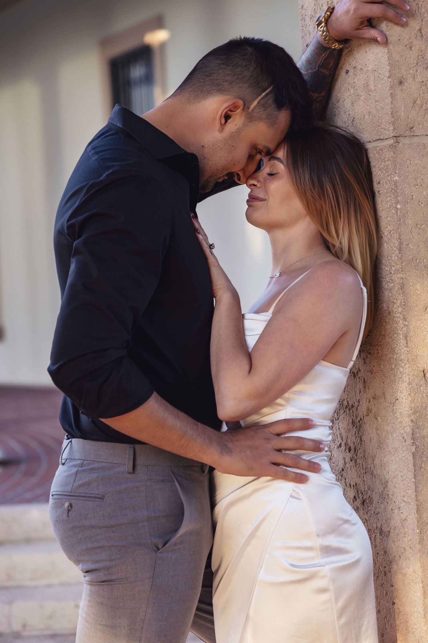 Romantic couple sharing an intimate moment against a stone wall in soft lighting.