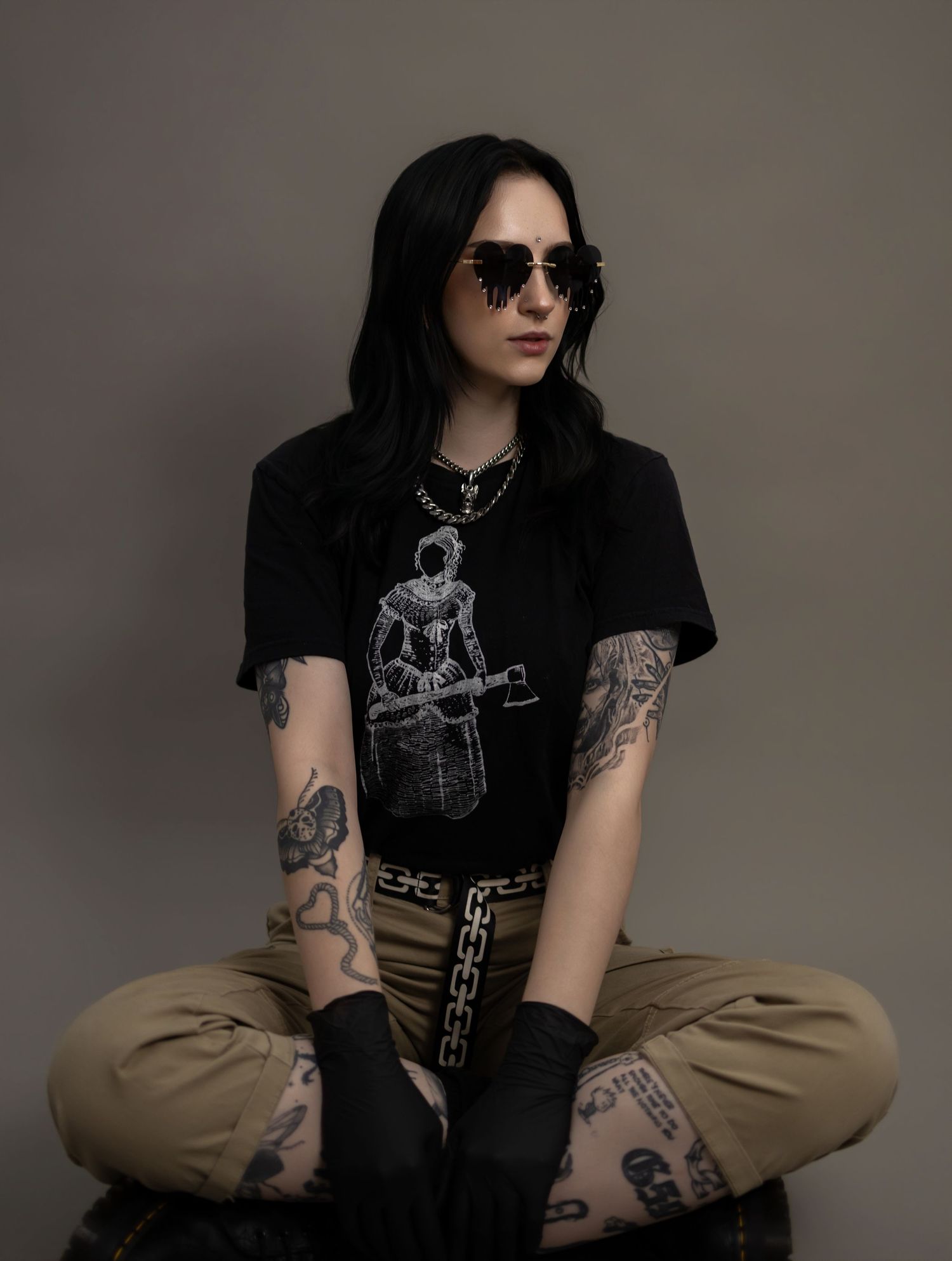 Tattooed woman in sunglasses sitting cross-legged, wearing a black graphic tee and cargo pants with extensive arm tattoos.