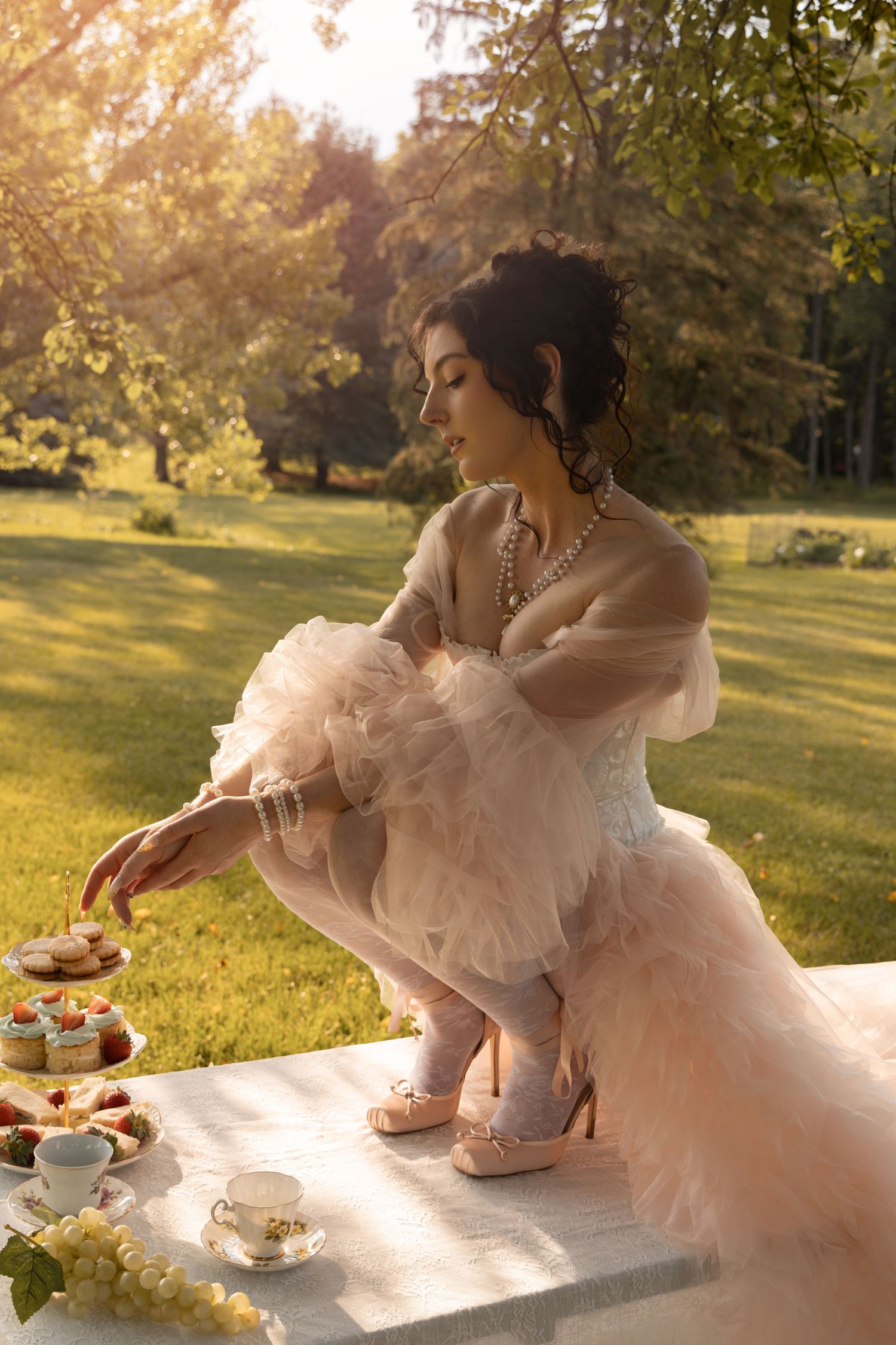 A woman in a pink tulle gown reaches for cupcakes at an outdoor tea party setup in a sunlit garden.