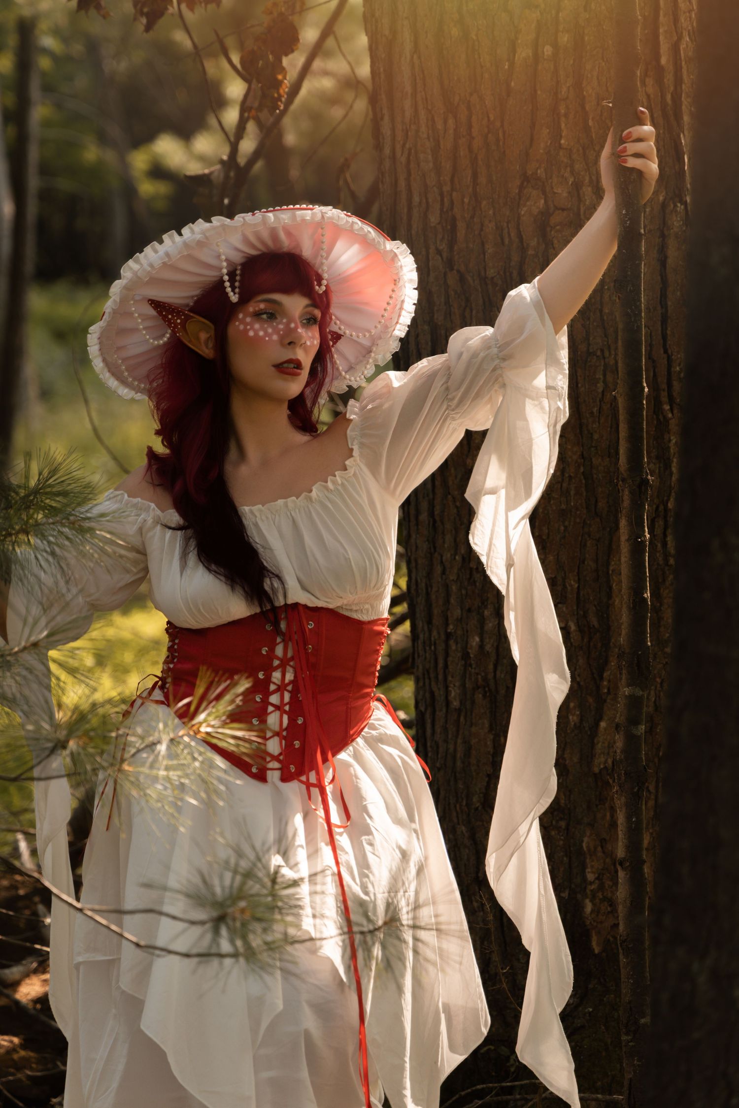 Woman in white Renaissance dress with red corset and pink hat poses dramatically against a tree in a sunlit forest.