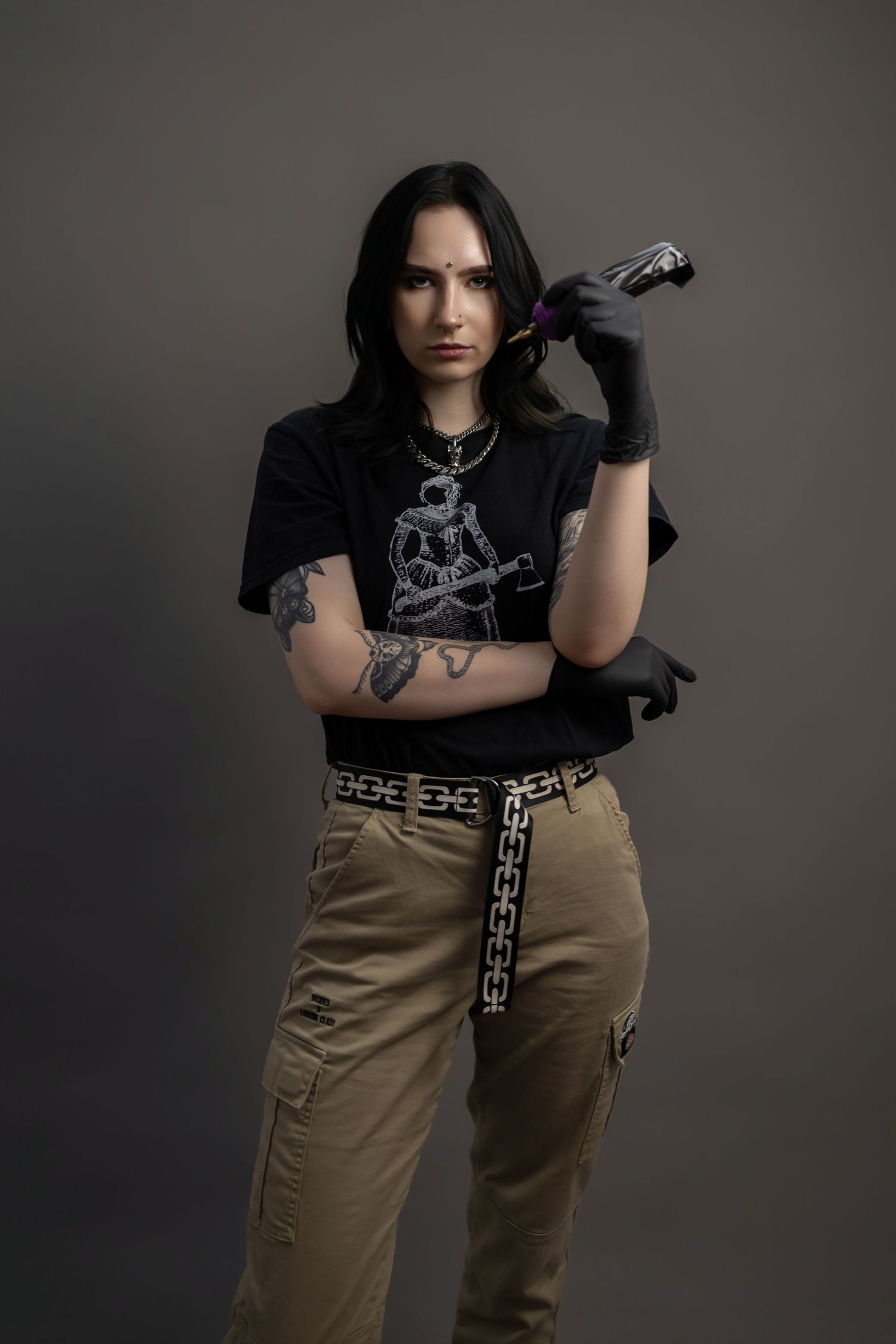 Tattooed female artist poses with tattoo machine in black gloves, graphic tee, and khaki cargo pants.