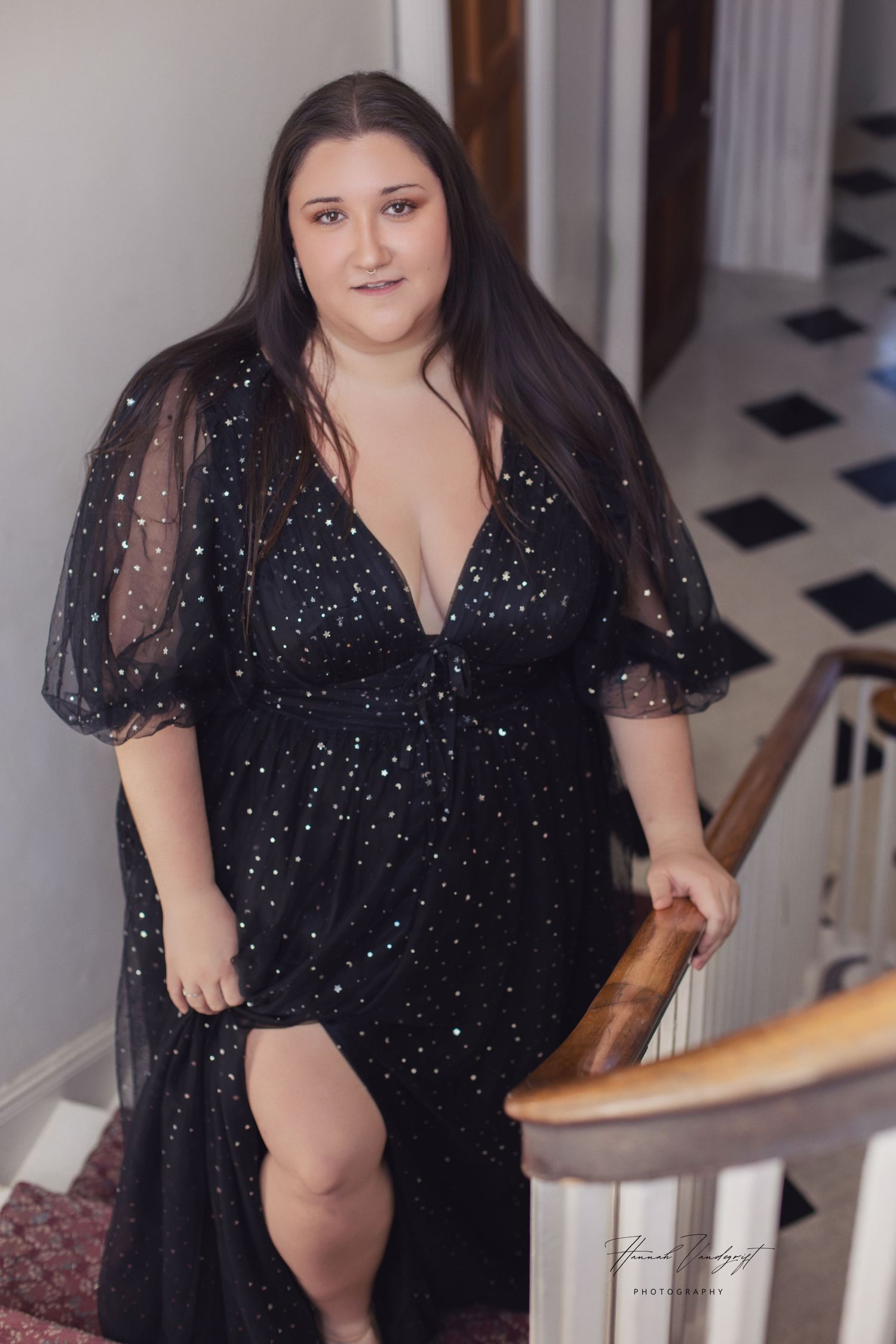 A figure in a sparkly black evening gown with sheer puff sleeves poses on stairs near a wooden banister.