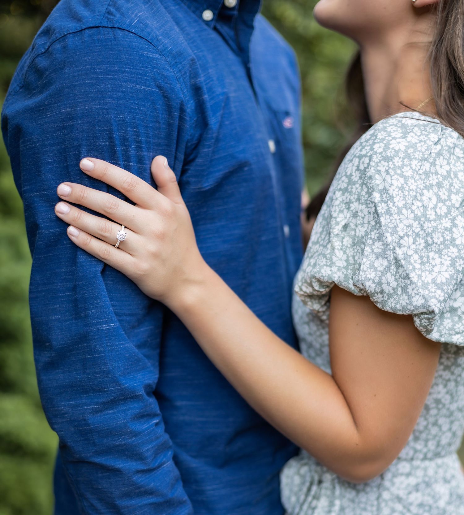 Close up of a couple's embrace showing a sparkling engagement ring on her hand against his royal blue button-down shirt.