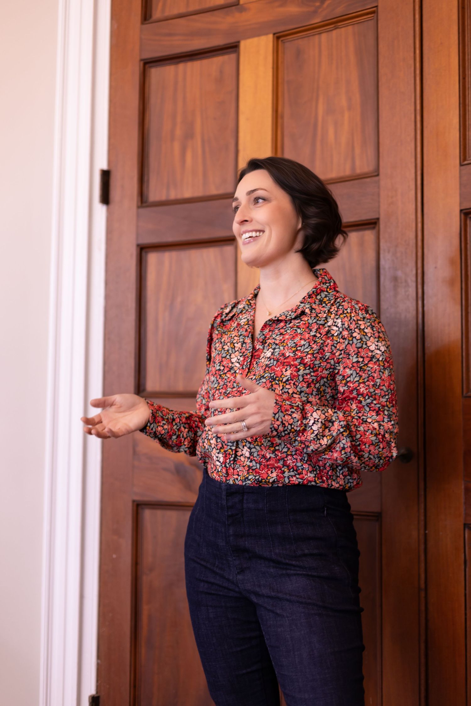 Woman in red floral shirt poses cheerfully by wooden door.