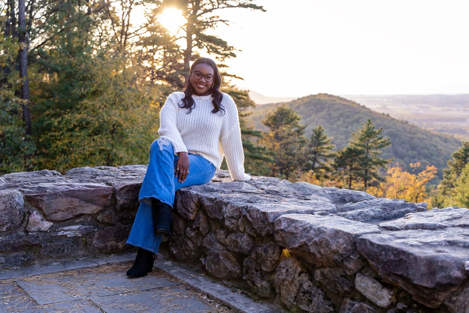 Model in white sweater and jeans sits on stone wall at scenic mountain overlook during sunset.