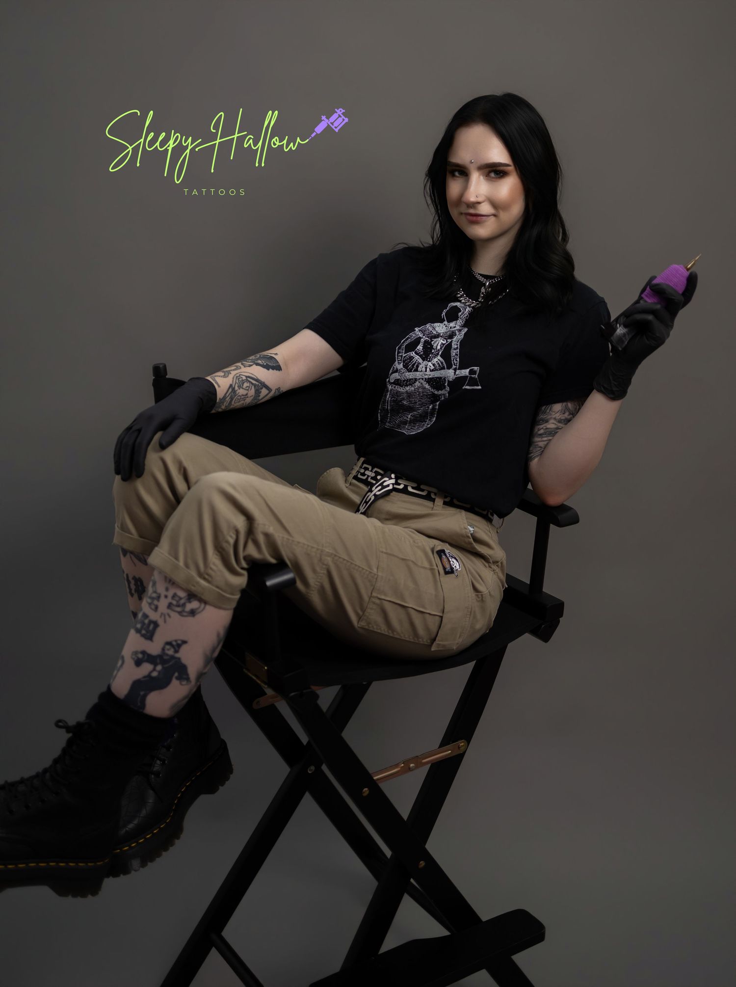 Tattooed female tattoo artist sitting in director's chair holding purple tattoo machine, wearing black tee and cargo pants.