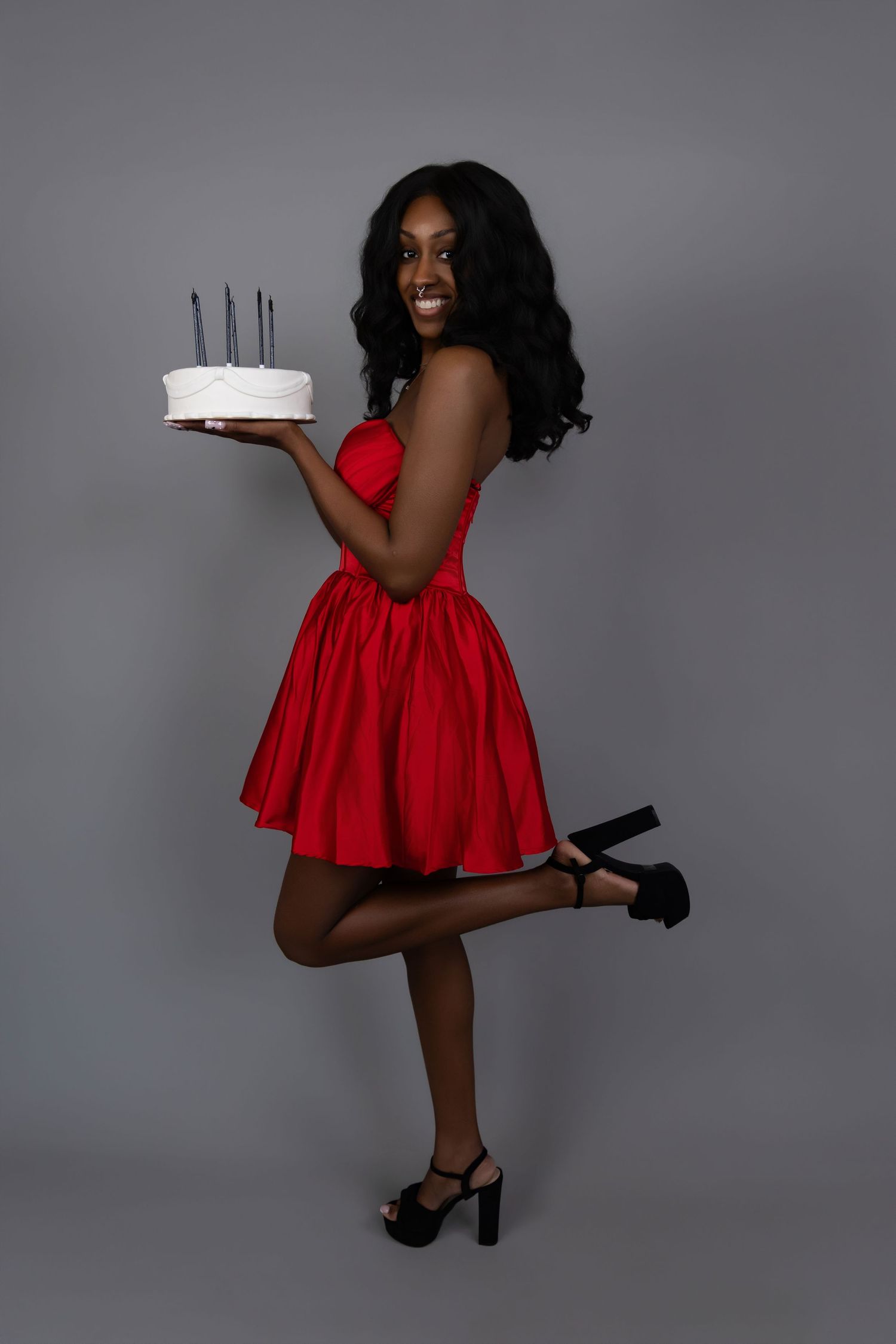 Smiling woman in red dress holding birthday cake with candles, posing on one leg against gray background.