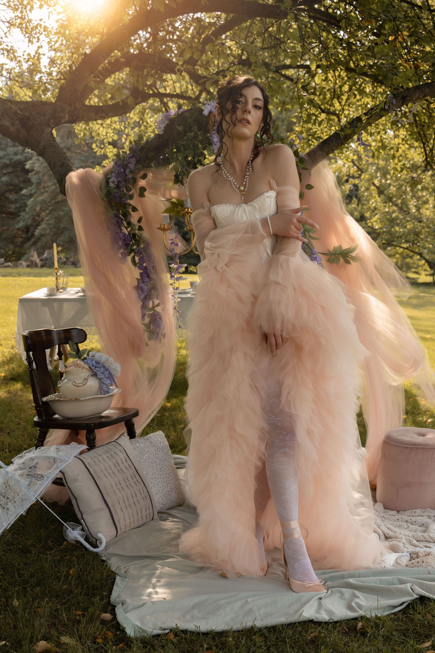 Woman in blush pink tulle gown poses outdoors under tree at golden hour with vintage decor.