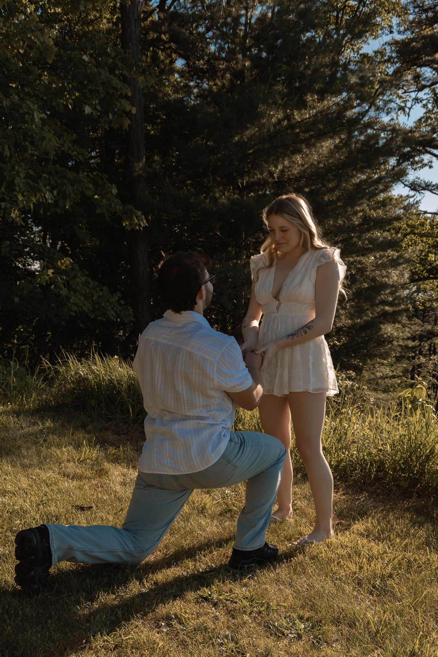 Man proposing on one knee to woman in white dress outdoors in sunlit grassy field with trees.