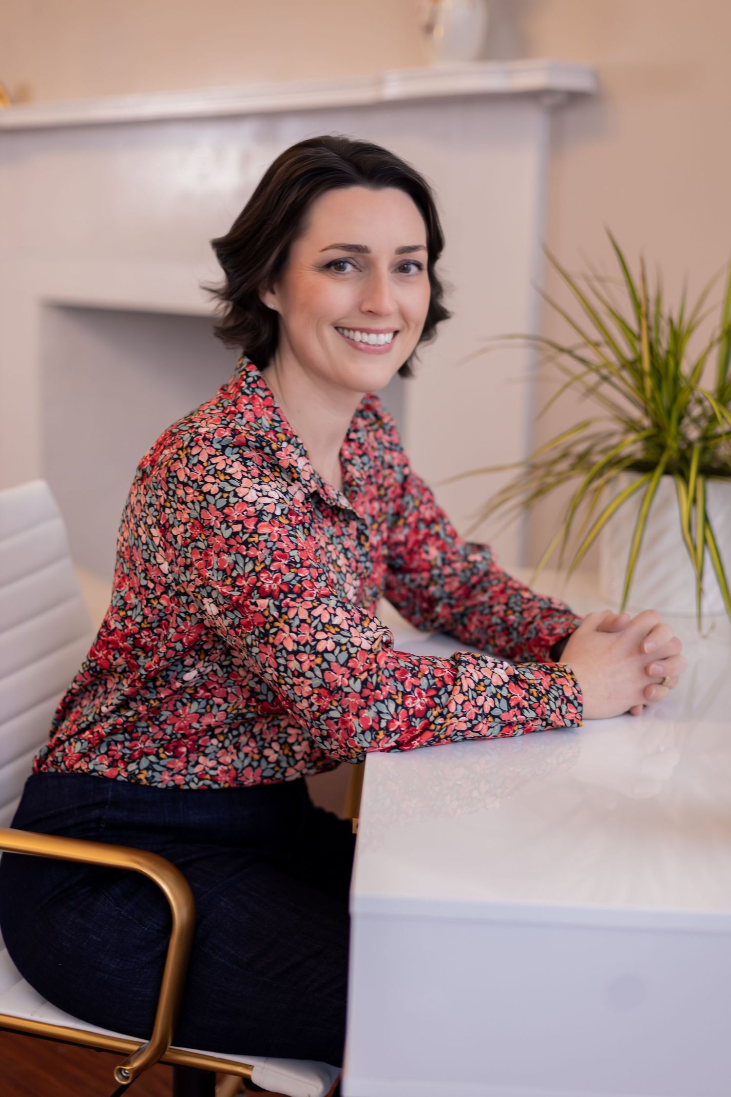 Business professional in floral blouse sits at white desk with warm smile.