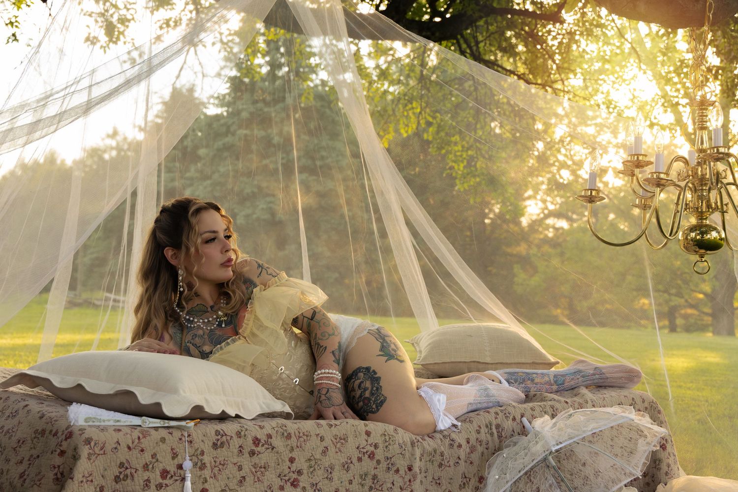 Tattooed woman reclining on floral outdoor daybed under sheer canopy with golden sunset light filtering through trees.