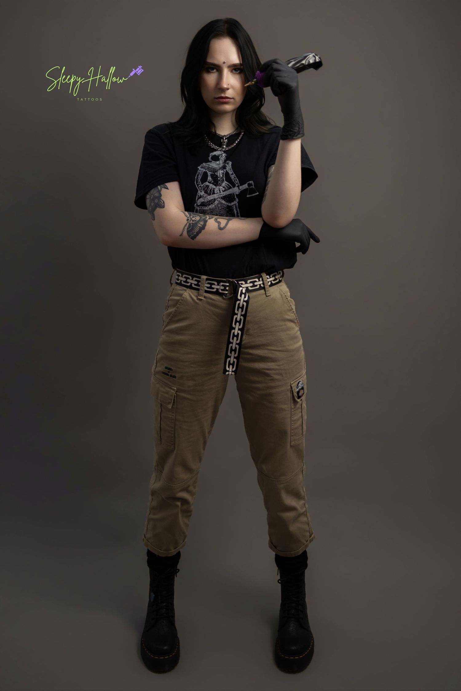 Tattooed woman in black graphic tee, cargo pants, and combat boots poses confidently holding a tattoo machine.