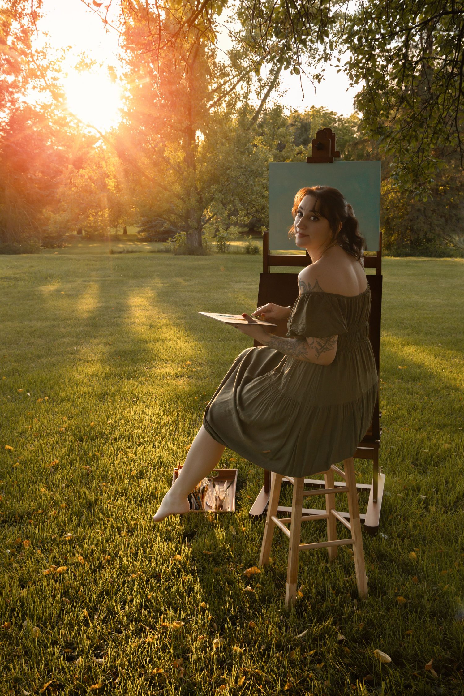 Woman in gray dress painting on easel outdoors at golden sunset in lush green park.