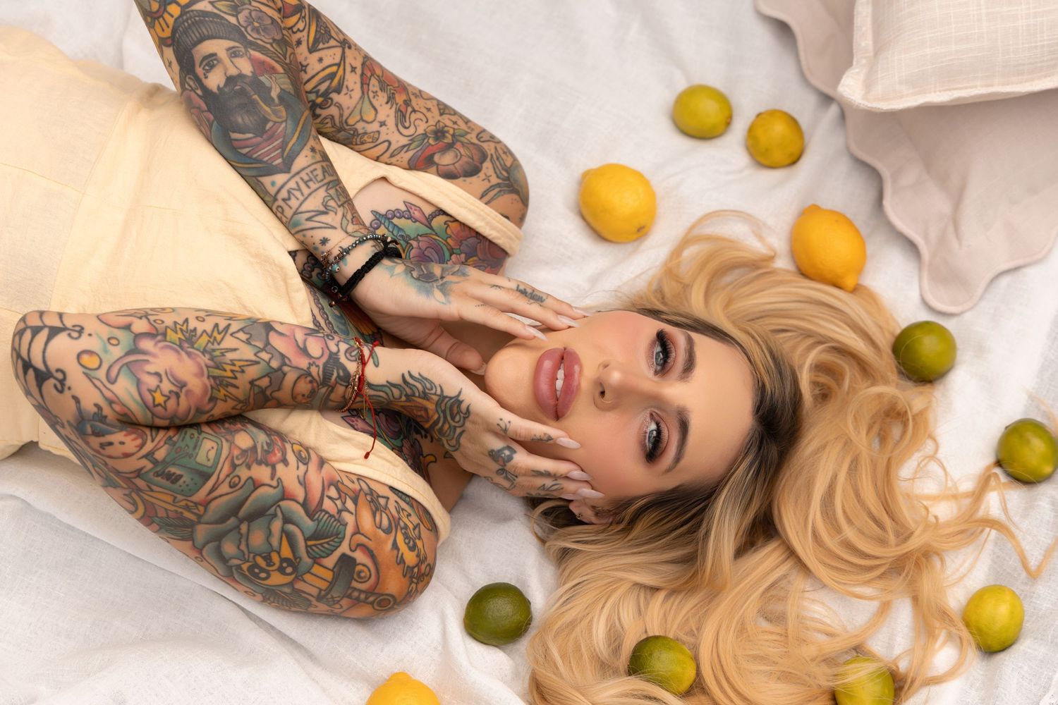 Tattooed blonde woman lying on white bedding surrounded by lemons and limes, wearing a cream dress.