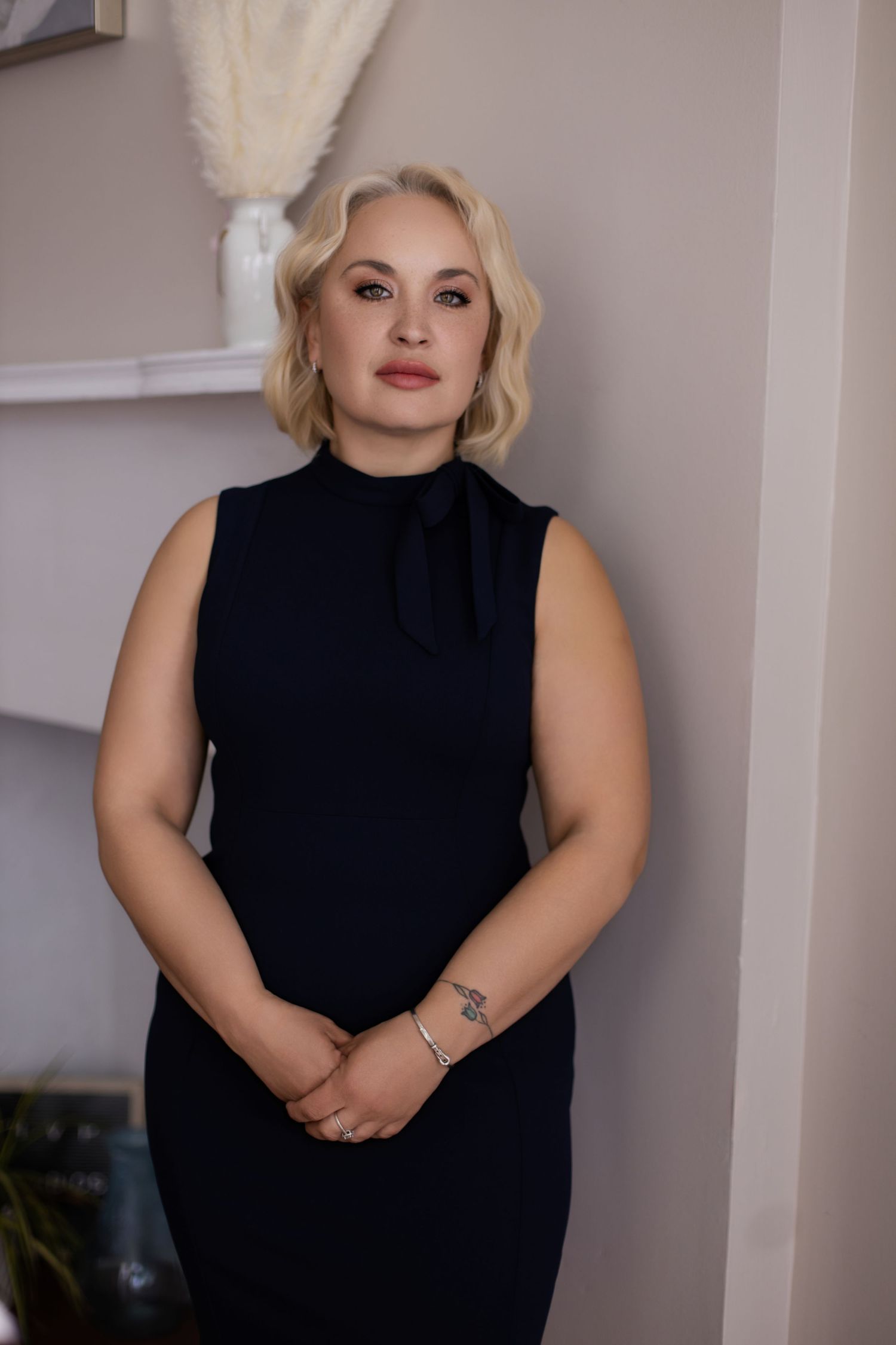 Portrait in navy sleeveless dress standing against neutral wall with decorative curtain.
