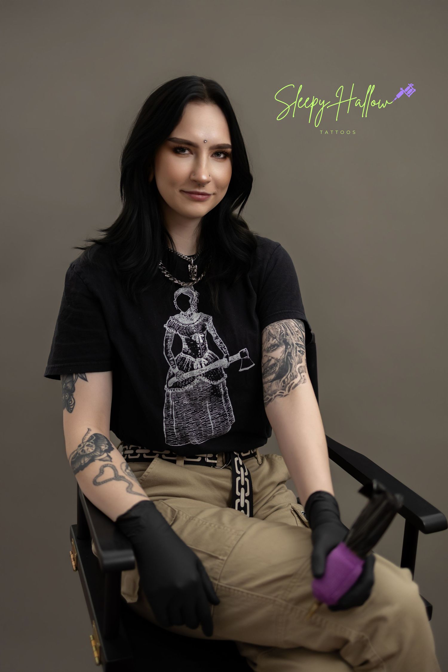 Tattooed female tattoo artist smiling in black graphic tee, khaki pants, and black gloves holding tattoo machine.
