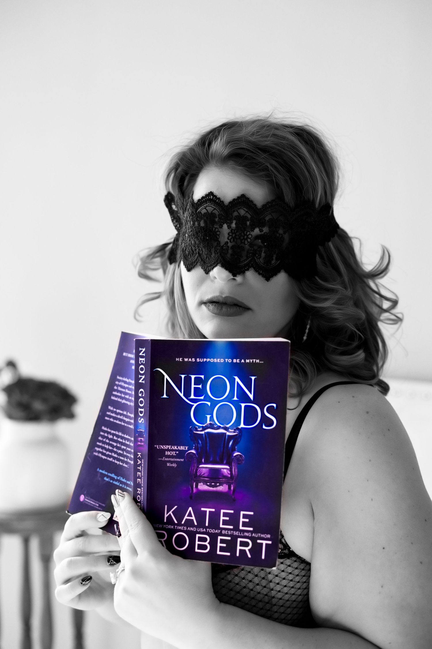 Model wearing lace eye mask holds Neon Gods book in artistic black and white photo.
