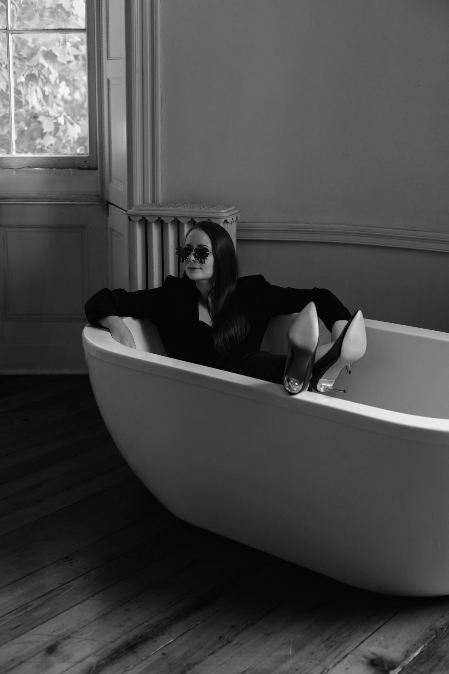 Moody black and white photo of a modern freestanding bathtub on hardwood floors near a bright window.