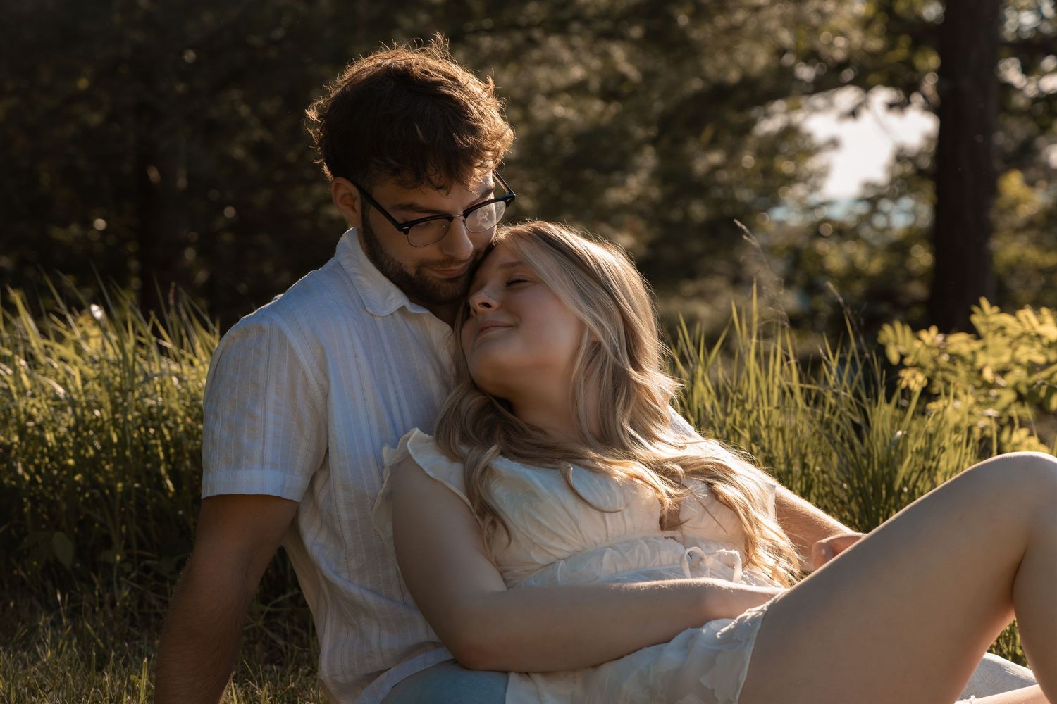 Romantic couple sitting together in sunlit meadow, man with glasses embracing smiling blonde woman outdoors.